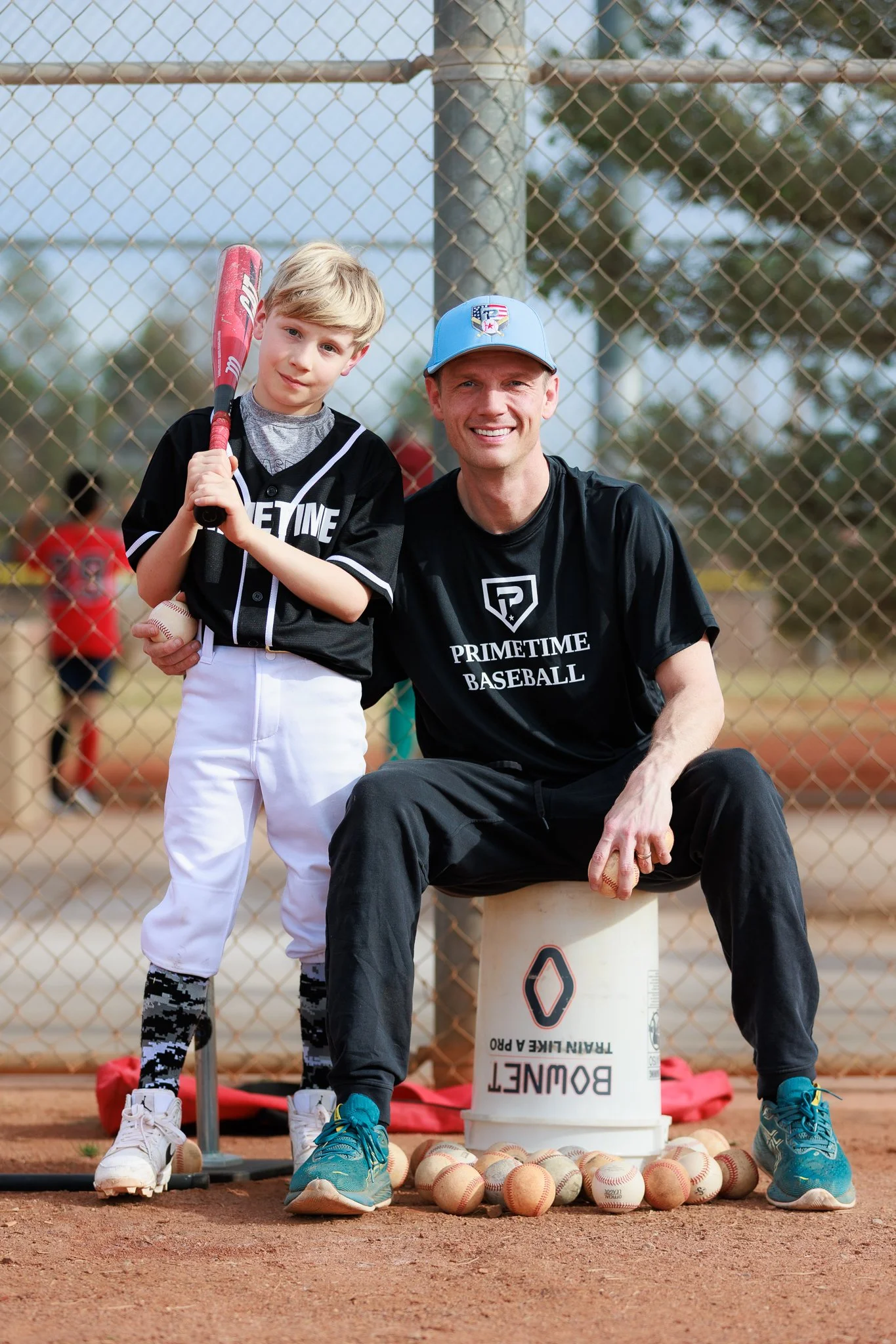 A boy and a man are on a baseball field behind a chain-link fence. The boy is holding a baseball bat and a baseball, and the man is sitting on a white bucket with baseballs around his feet. Both are smiling and wearing sports attire, with trees and a