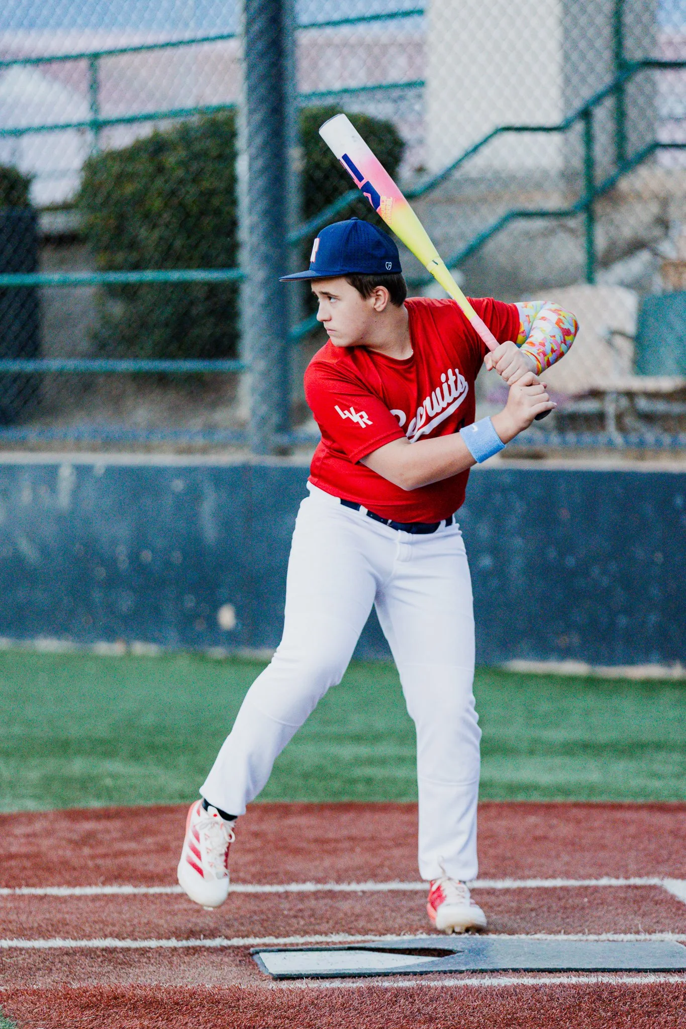 A young boy in a red baseball jersey and white pants practicing baseball batting on a field. He is holding a yellow and purple bat and is about to hit a baseball. He is wearing a navy blue cap and colorful arm sleeves, with white and red sports shoes
