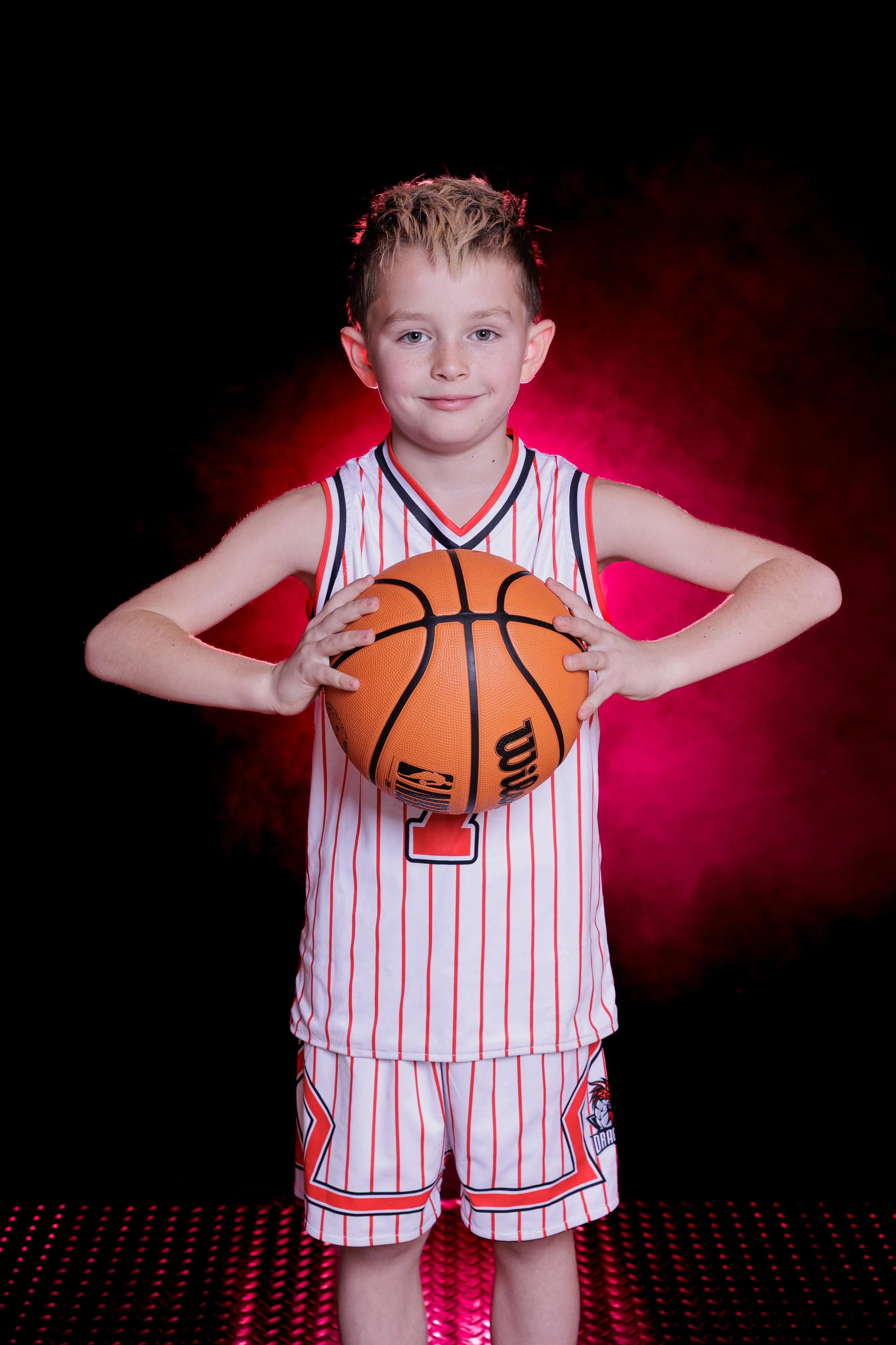 A young boy in a basketball uniform holding a basketball, standing on a pink-lit gaming mat, with a black background and red lighting around.