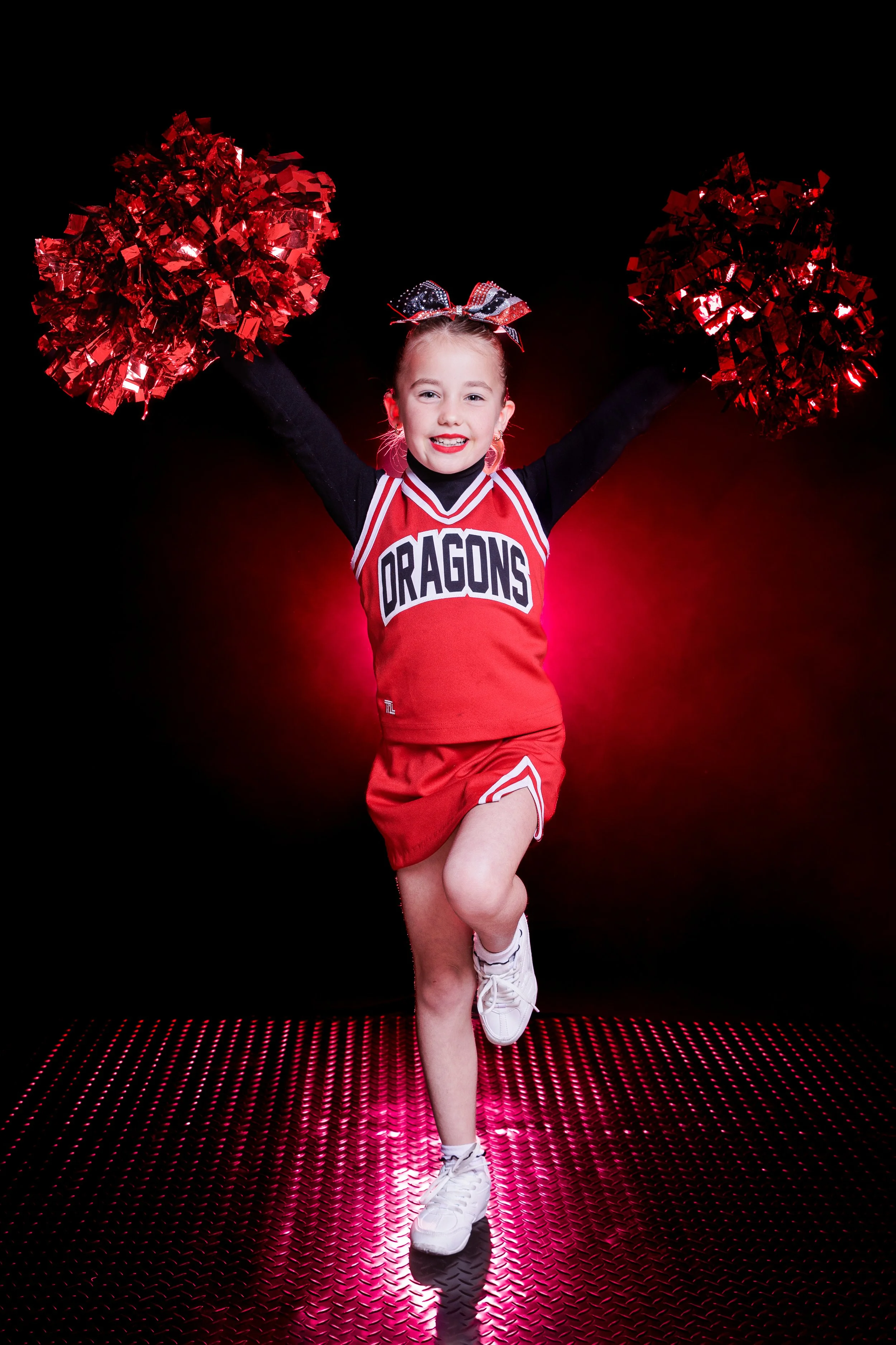 Young girl cheerleader in red and black uniform with 'DRAGONS' written on it, holding red pom-poms, smiling, standing on a pink illuminated floor with a black background.