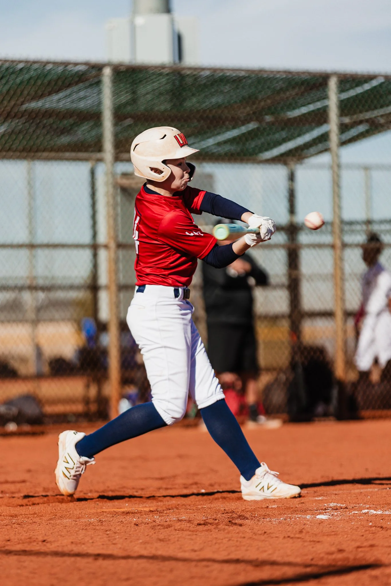 A young baseball player in a red uniform and helmet swinging a bat at a ball on a dirt field, with a chain-link fence and spectators in the background.