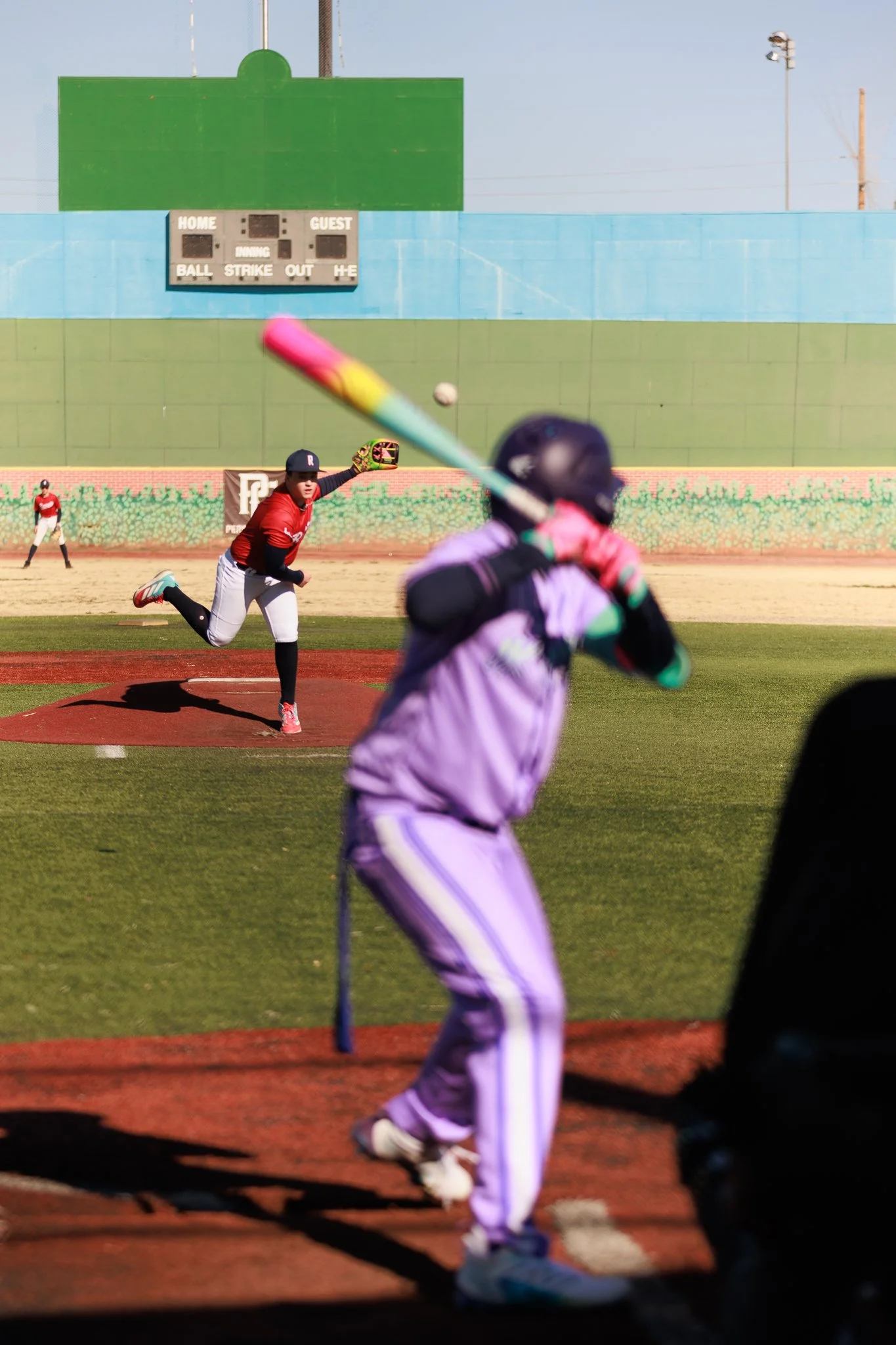 A baseball game is in progress, with a pitcher in a red jersey throwing the ball towards a batter in purple and green protective gear. Several players are visible in the background, and a scoreboard can be seen on a green wall behind the players.