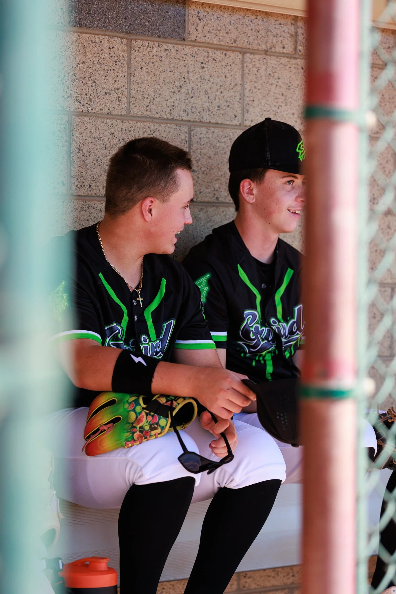 Two young men sitting against a brick wall, wearing baseball uniforms, with one smiling and the other looking down. They are partially obscured by a fence in the foreground.