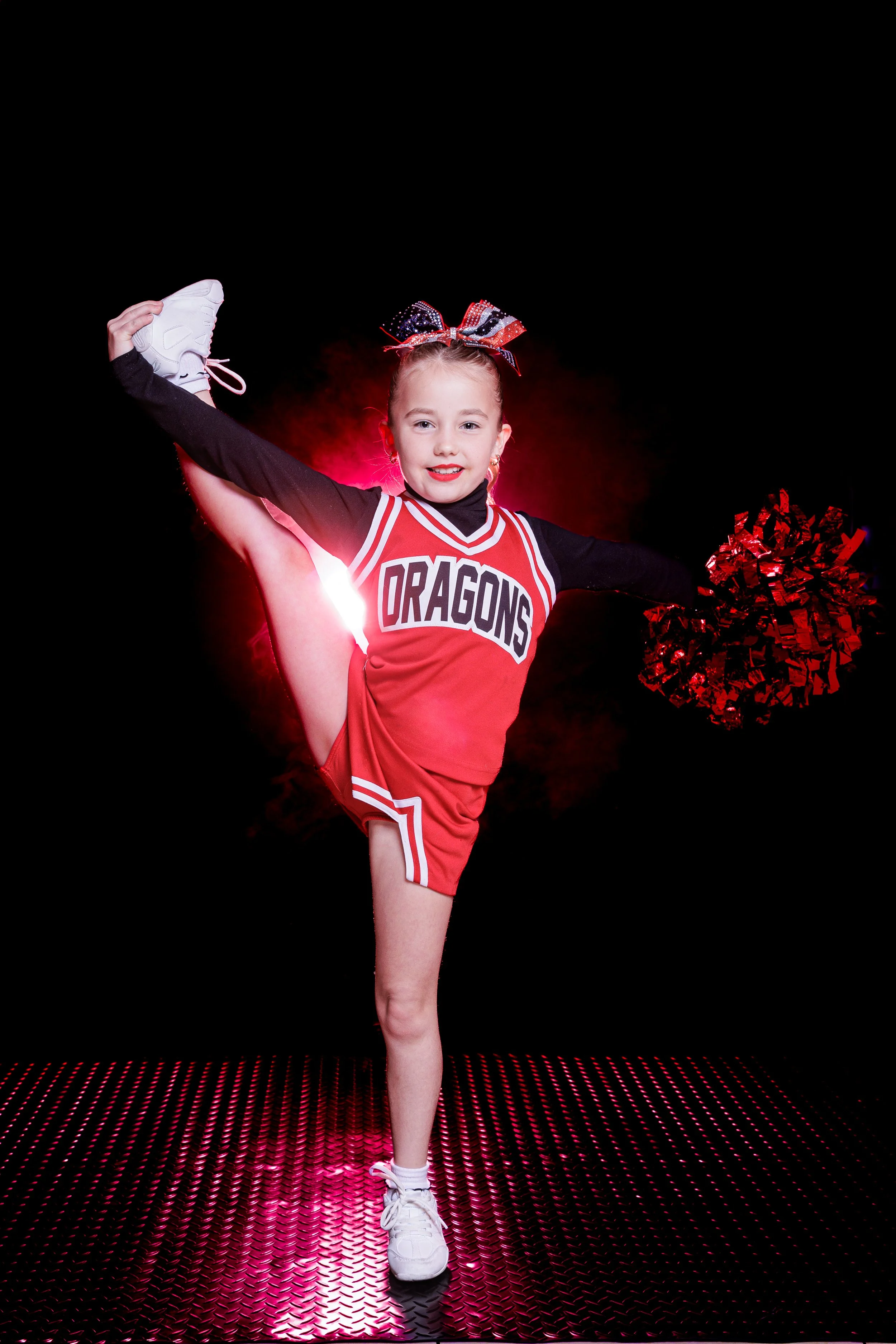 A young cheerleader in a red and black cheerleading uniform with "DRAGONS" on it performing a high kick while holding red pom-poms. She is standing on a reflective, textured surface with a dark background.