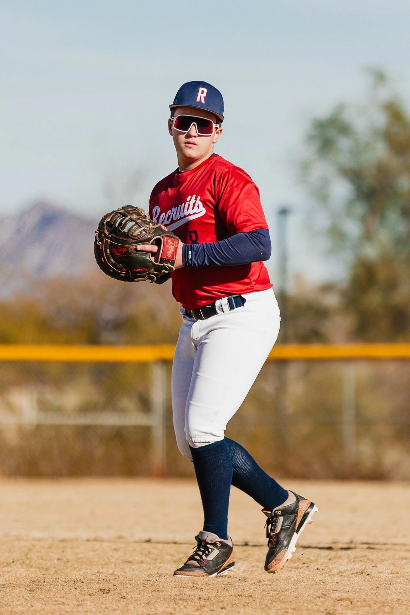 A baseball player in a red jersey and white pants standing on the field, wearing sunglasses, a cap, and black cleats, holding a baseball glove.