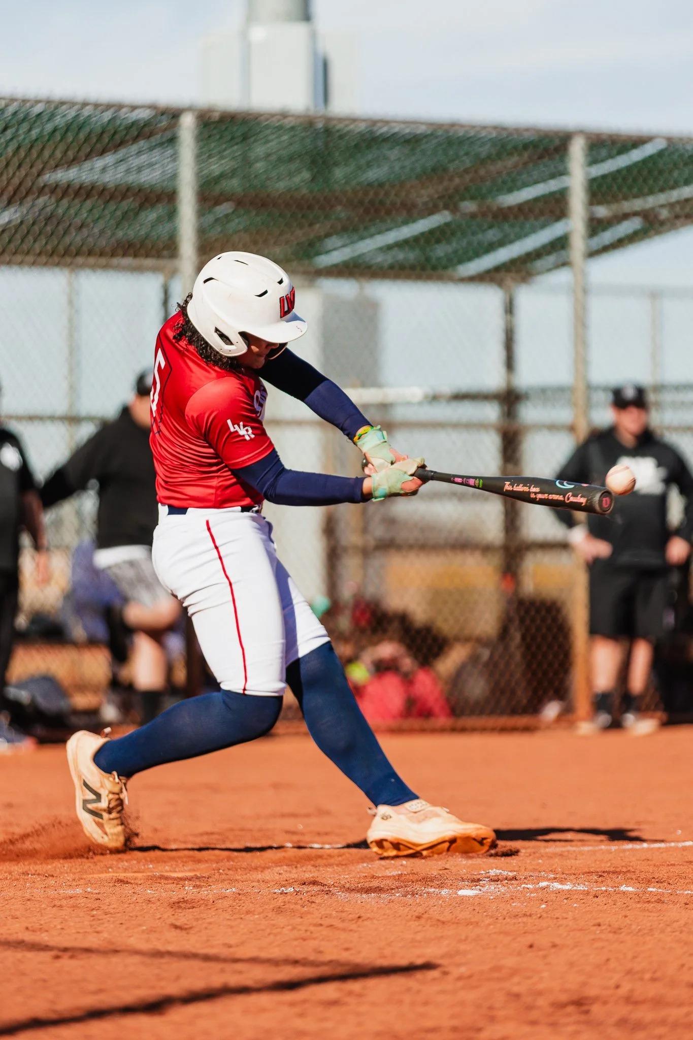 A female softball player in a red jersey and white pants is swinging a bat on a dirt field. She is wearing a white helmet and blue socks, with spectators and a fence in the background.