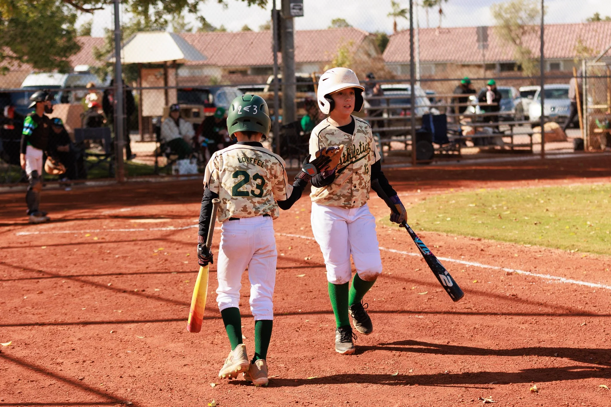 Two young baseball players dressed in camouflage uniforms and helmets on a baseball field. One player is holding a bat, and the other is holding a bat and appears to be talking. Background shows a chain-link fence, spectators sitting on bleachers, an