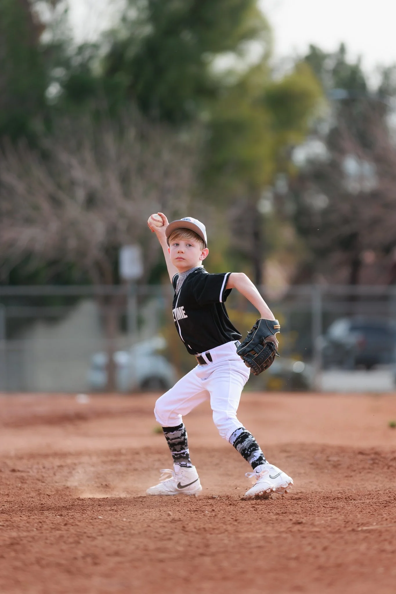 A young boy wearing a white baseball cap, black and white socks, white baseball pants, and a black shirt is pitching a baseball on a baseball field. The background shows trees and a fence.