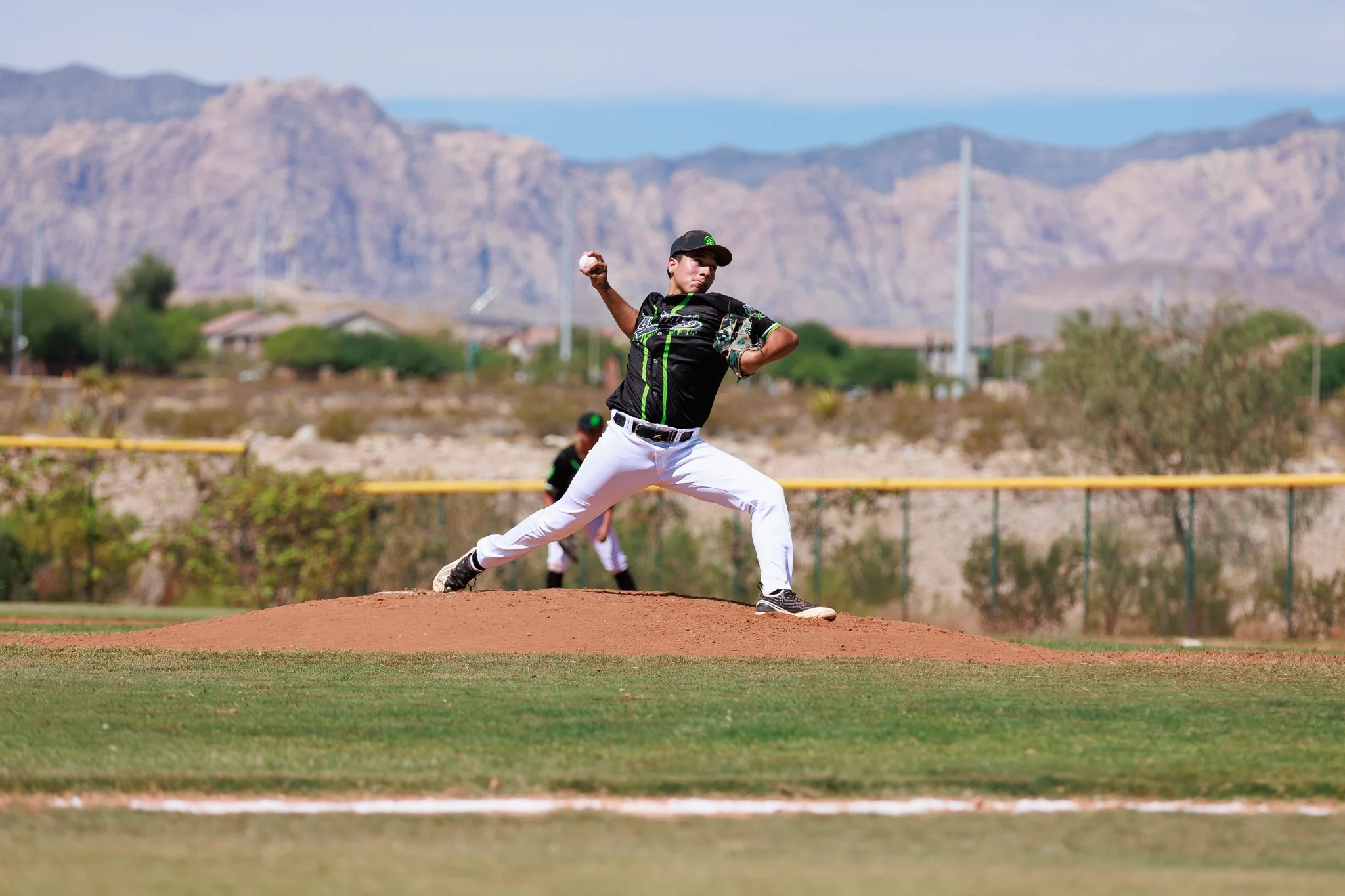 A young male baseball pitcher wearing a black and green uniform and a black cap is pitching on a baseball field with mountains in the background.