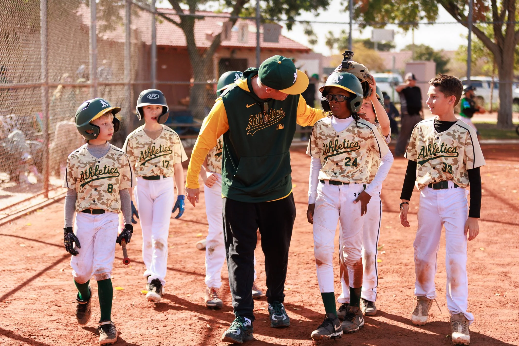 Little league baseball players walking with their coach on a baseball field with trees and a chain-link fence in the background.