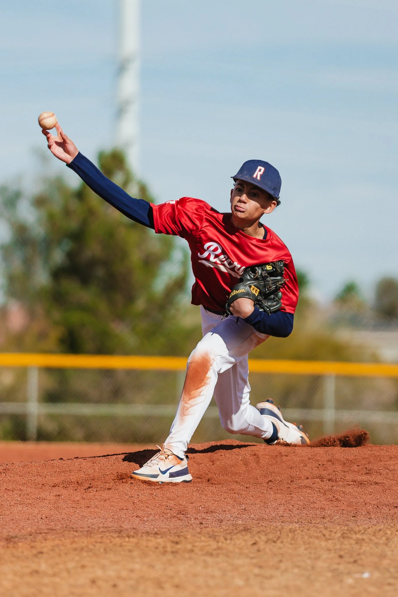 A young baseball player in a red uniform and dark blue hat is pitching a baseball on a reddish clay field during daytime. The player is in a crouched pitching stance, releasing the ball with his right hand, with a focused expression.