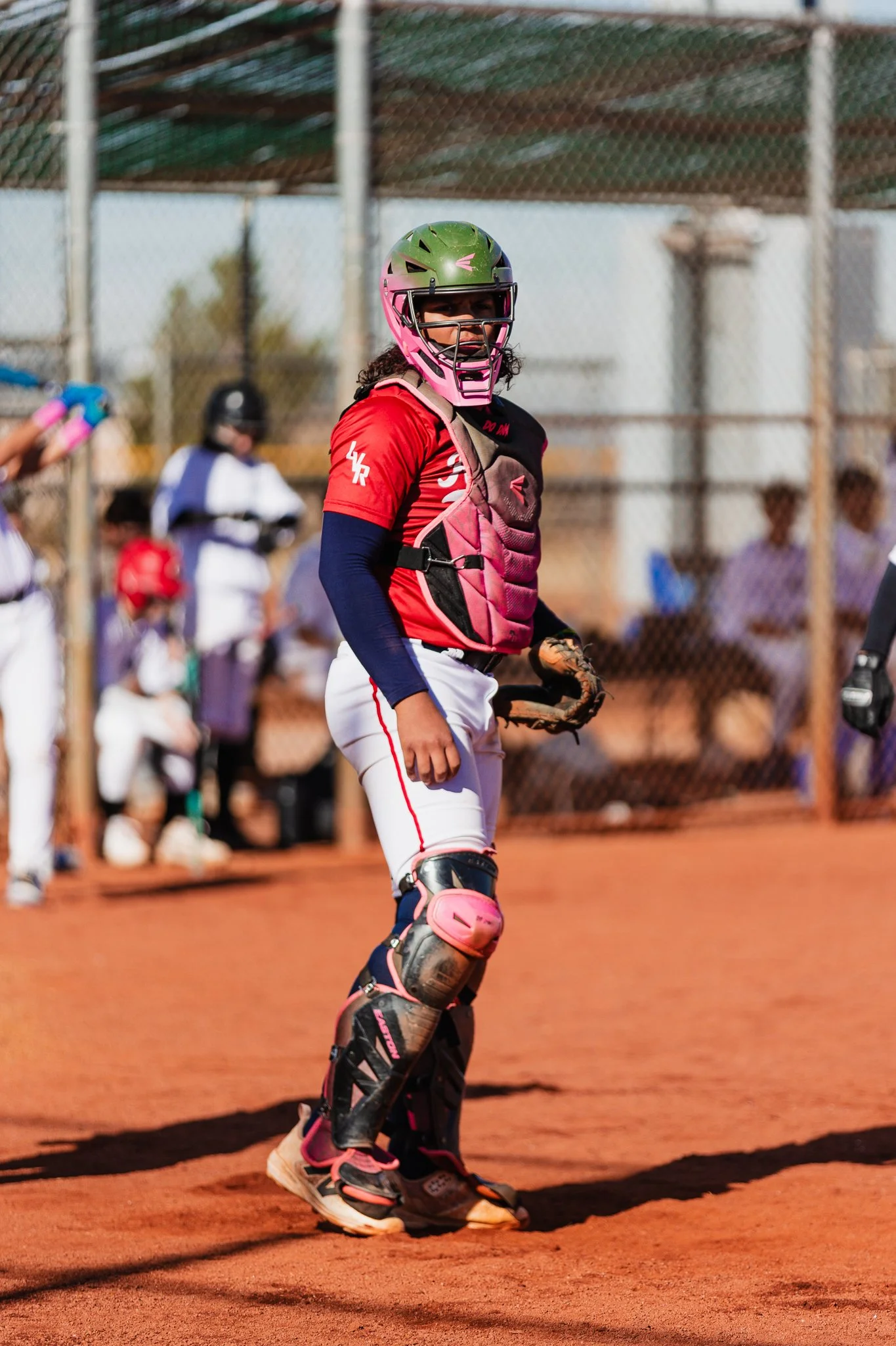 A female softball catcher in full gear, including a green helmet with a pink face mask, pink chest protector, and shin guards, standing on a dirt field during a game. There are other players and people blurred in the background behind a chain-link fe