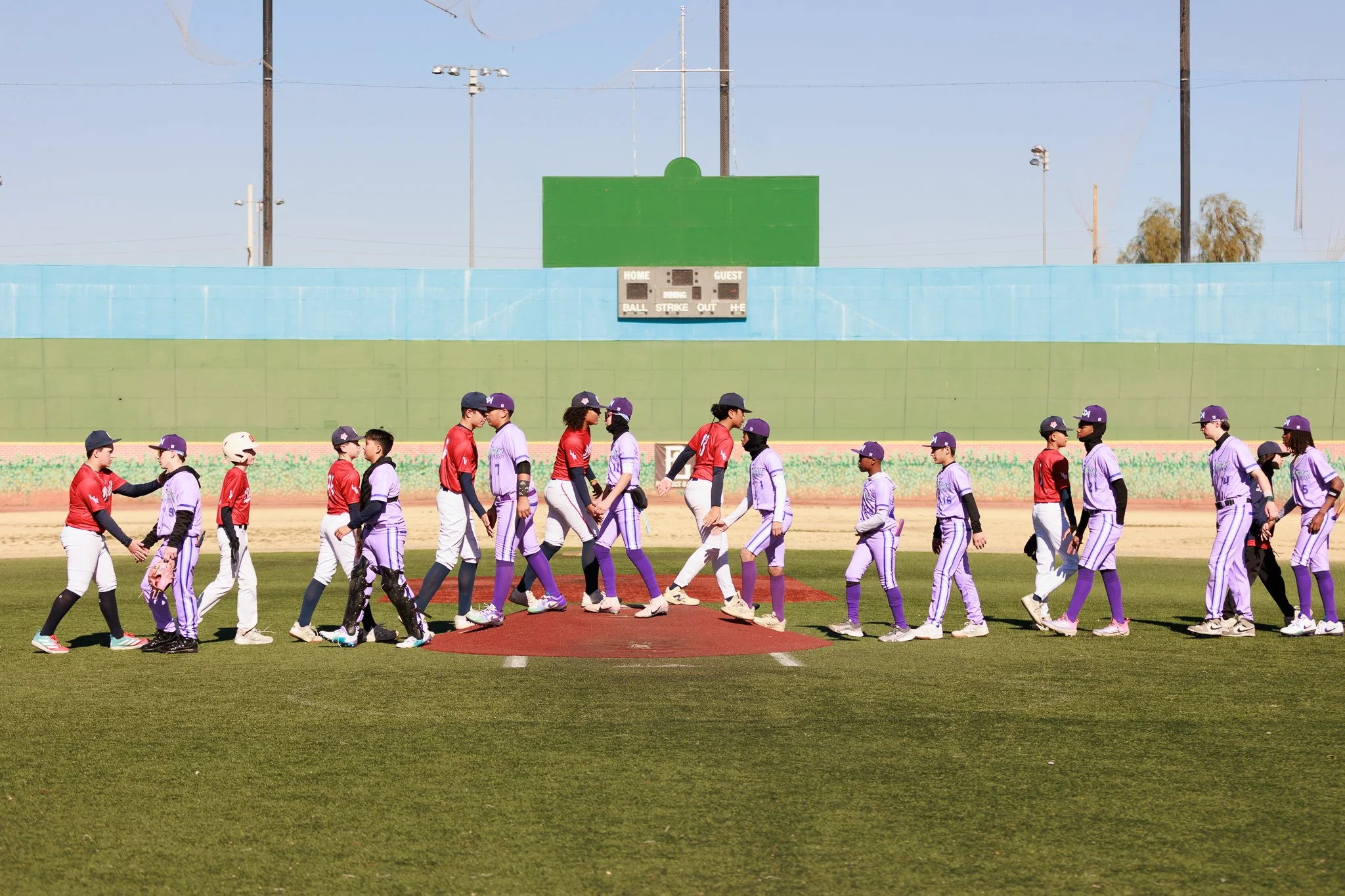 Young baseball players walking onto the field, shaking hands before the game, with a scoreboard and blue wall in background.