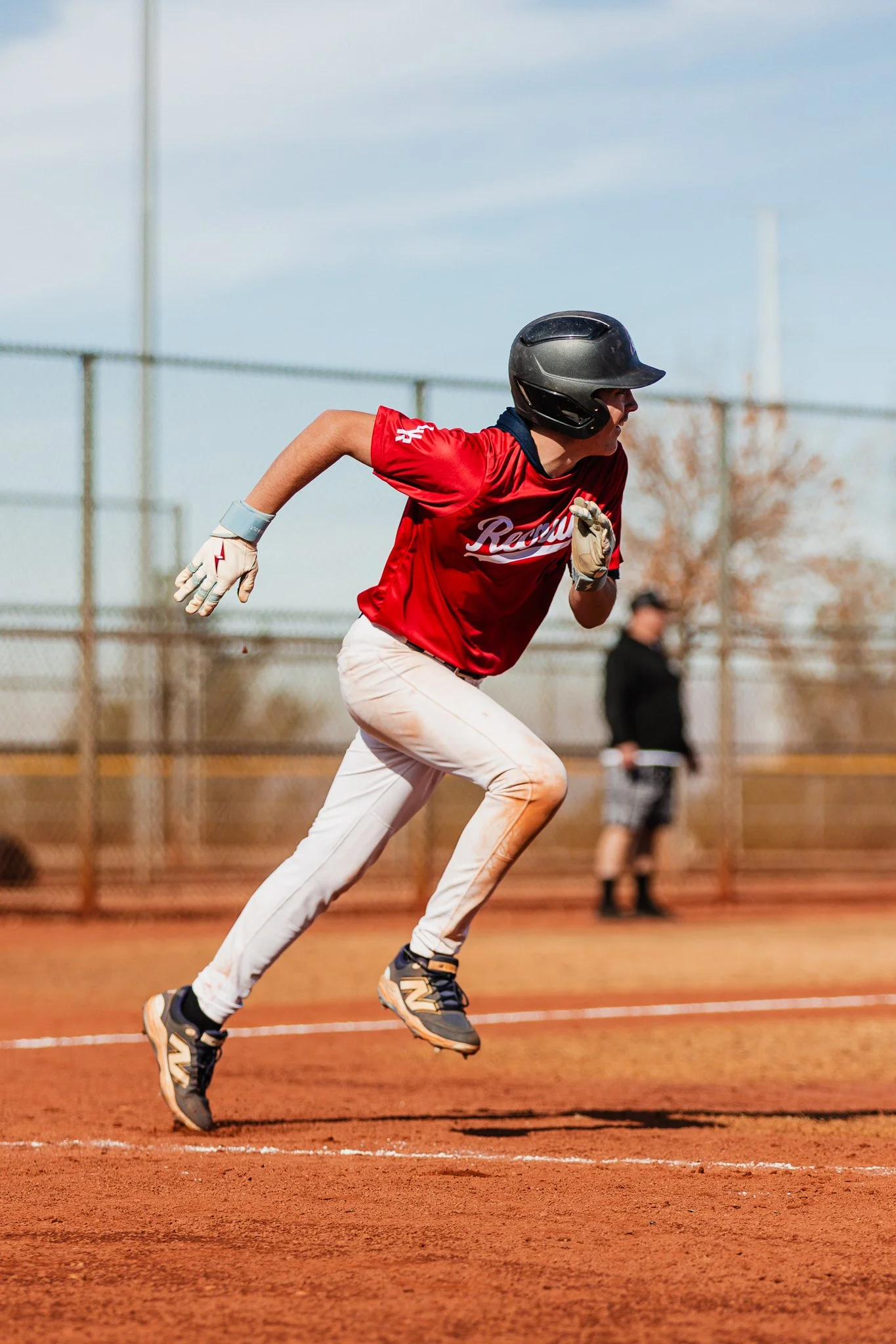 A baseball player running on a baseball field during daytime, wearing a black helmet, red jersey, white pants, and black cleats with yellow accents.