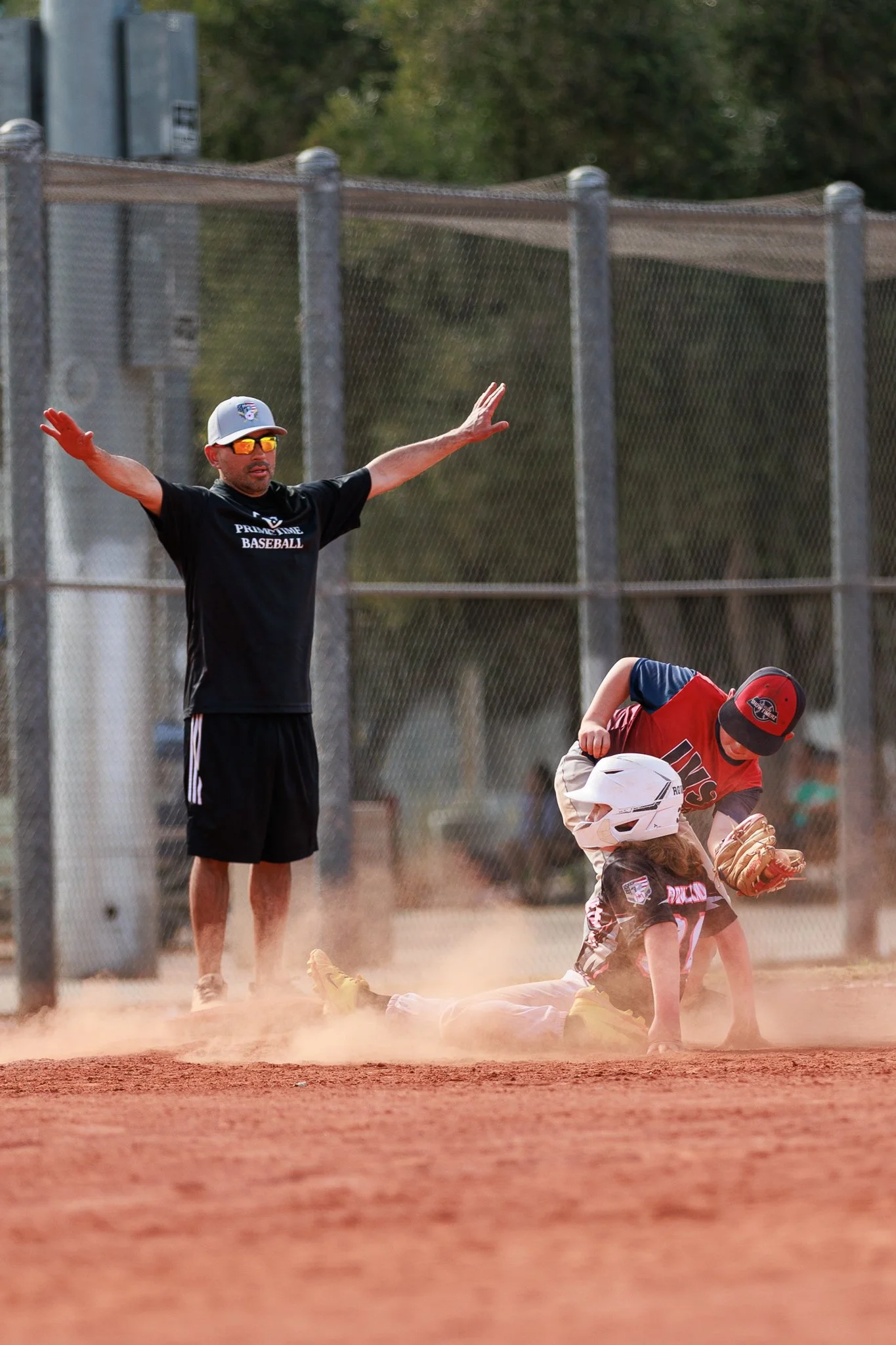 A youth baseball game with a coach and players on the field. The coach is standing with arms outstretched, wearing sunglasses and a cap. A player is sliding into a base while another player is preparing to catch the ball, date and time unknown.