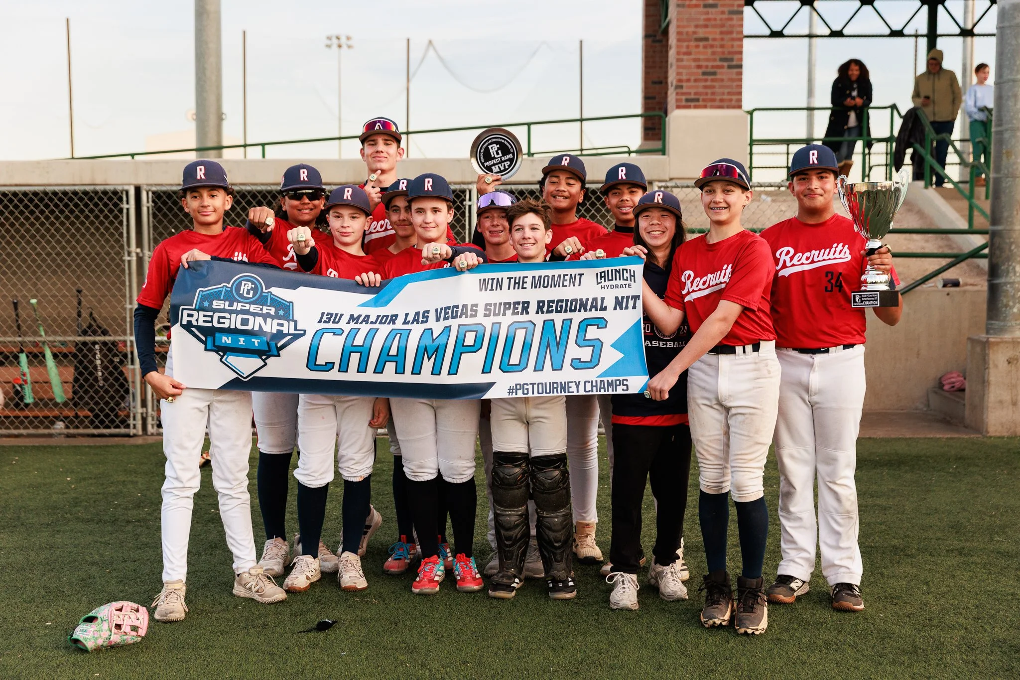 A youth baseball team celebrating winning a championship, holding a banner that reads 'Champions,' with some players wearing caps with the letter 'R' and one holding a trophy. The team is standing on a sports field with a chain-link fence and people 