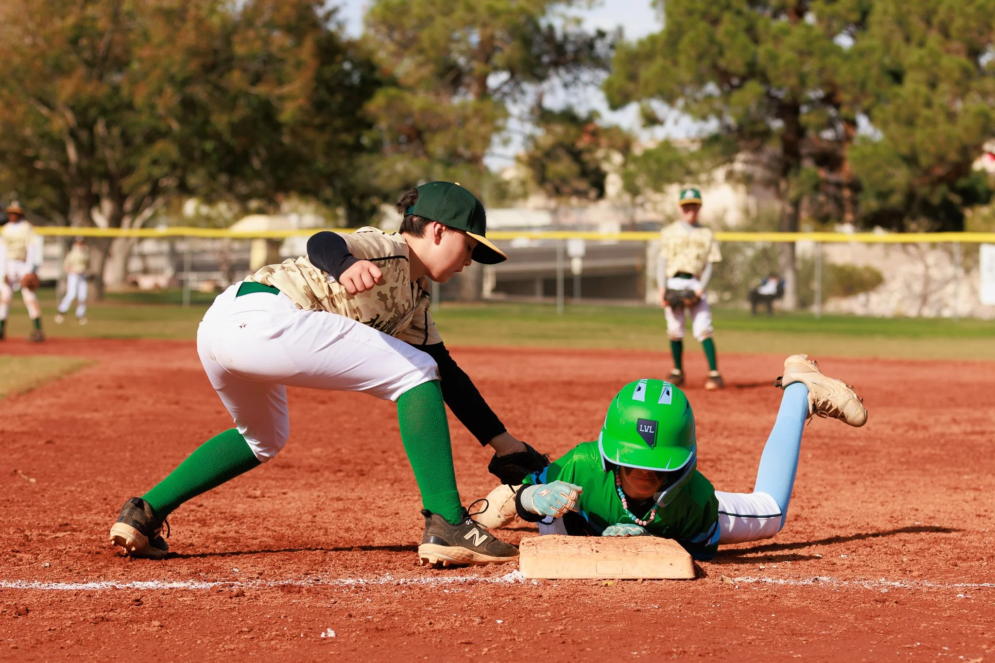 A young baseball player is sliding into second base while a teammate attempts to tag him out, with other players and trees visible in the background.