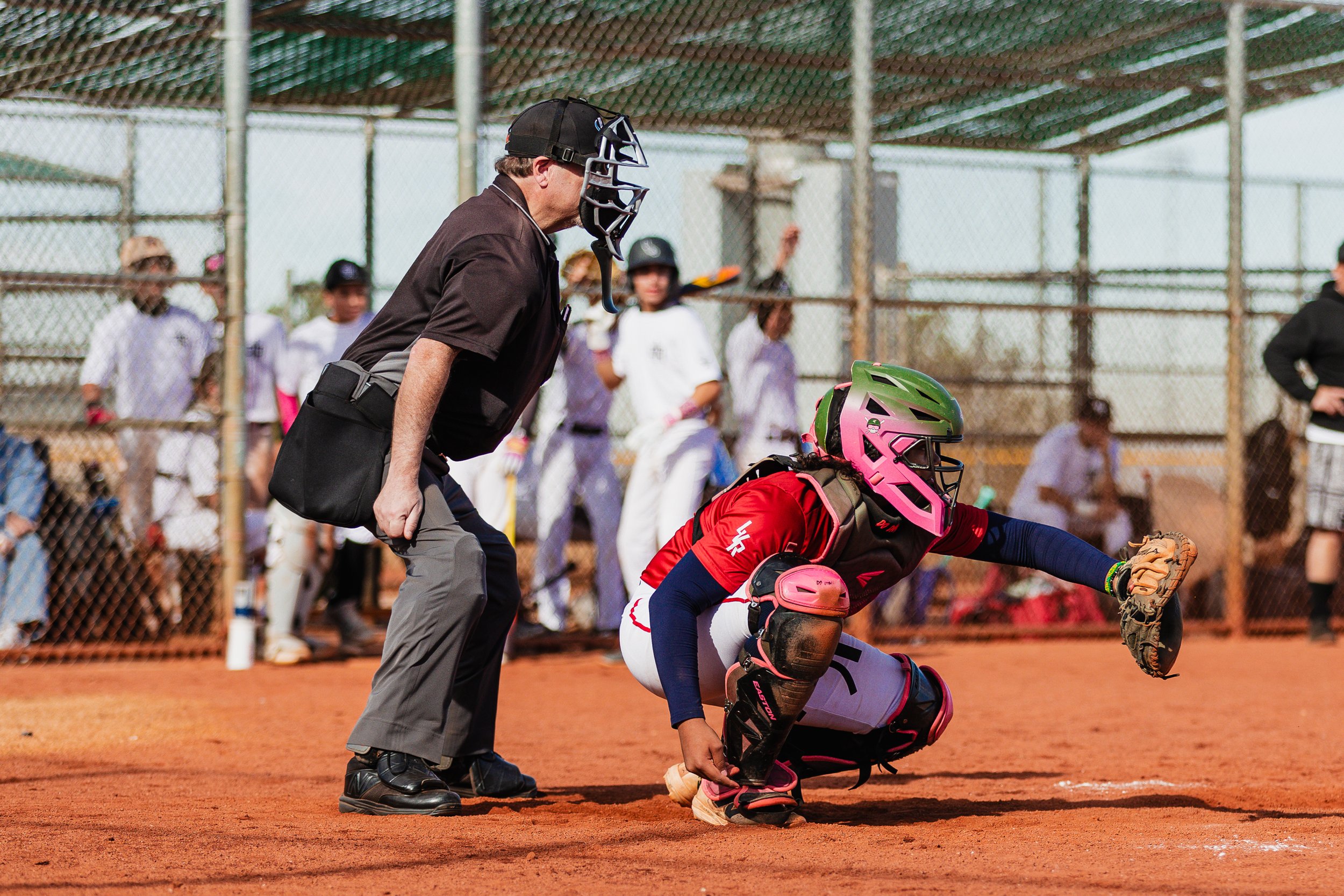 A baseball umpire wearing a black mask and black shirt stands behind a woman catcher in pink helmet and uniform on a baseball field, with a chain link fence and spectators in the background.