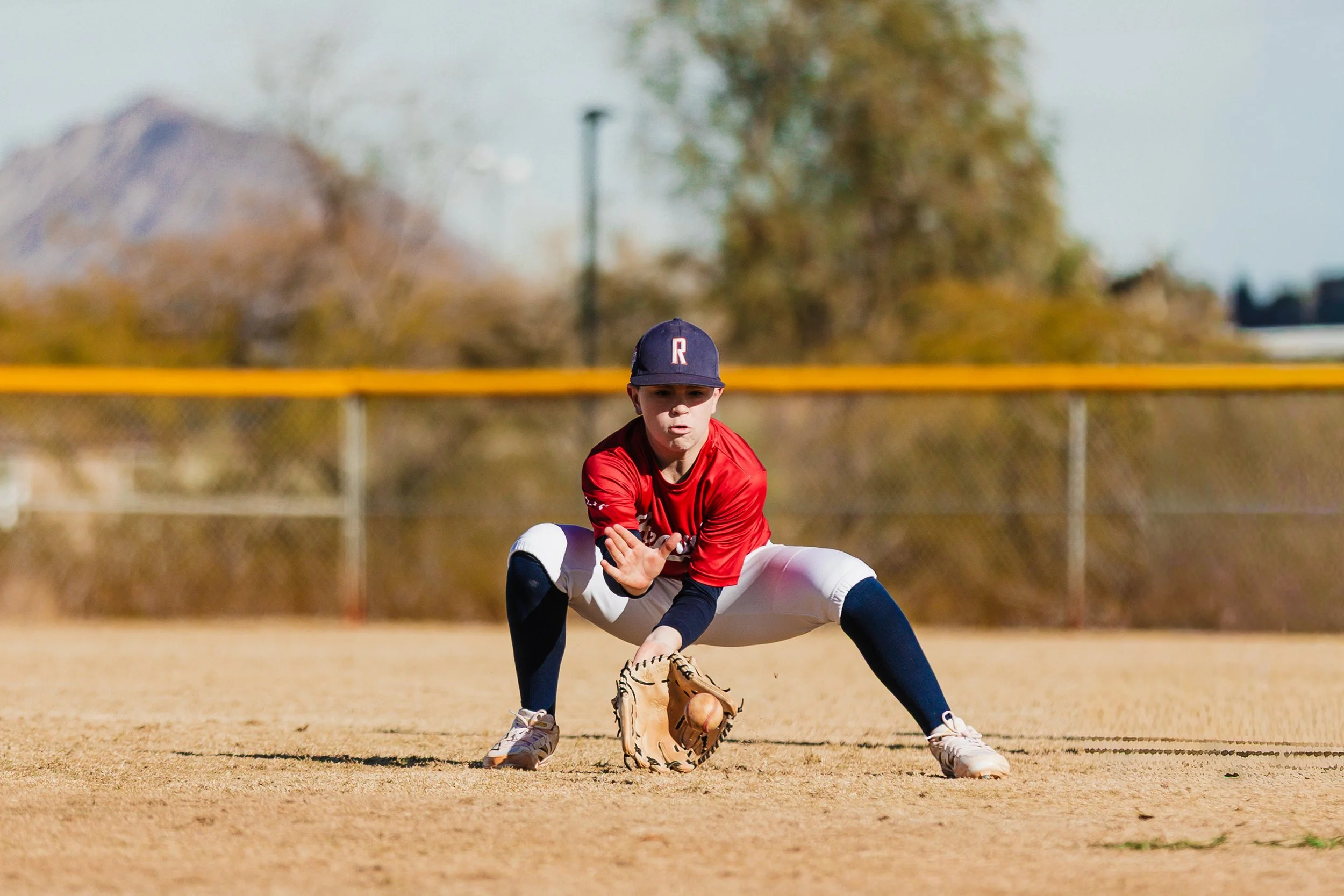 A young baseball player in a red jersey, black cap with a red and white letter, and white pants is fielding a ground ball on a dirt baseball field, with a chain-link fence and trees in the background.