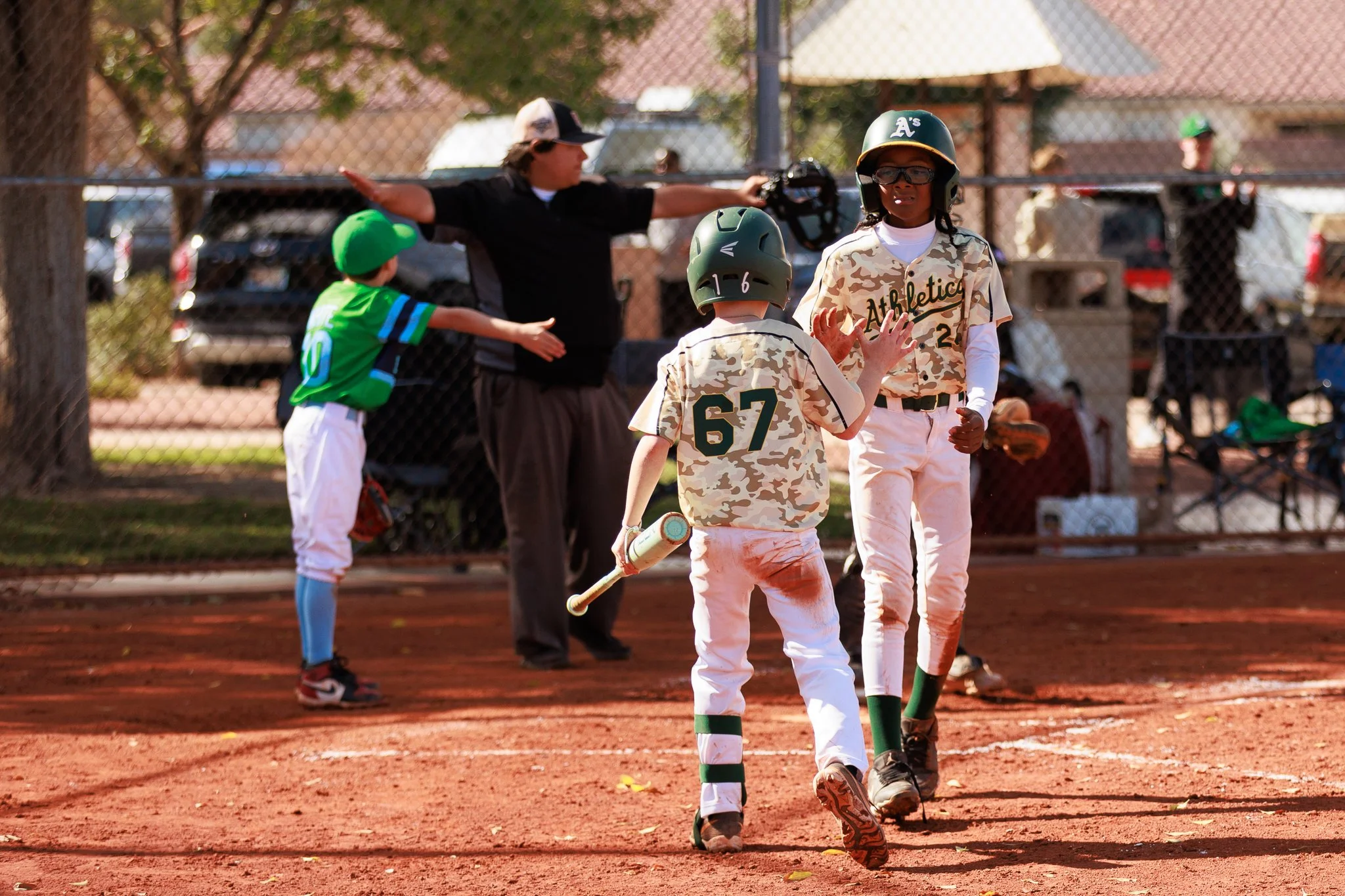 Young baseball players in camouflage jerseys and helmets at a baseball field, with coach and spectators in the background.