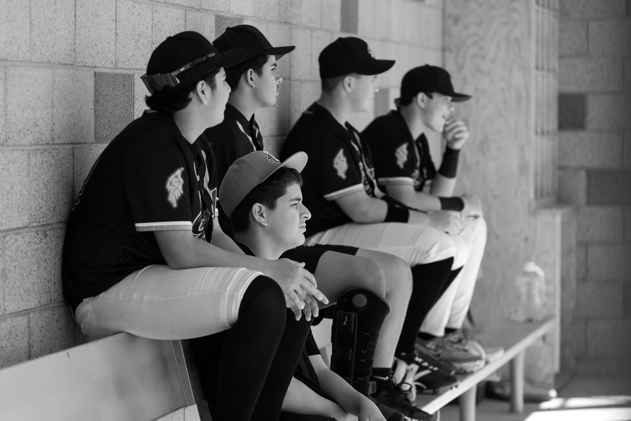 A black and white photo of five young male hockey players sitting on a bench against a brick wall. They are wearing hockey jerseys, caps, and some have hockey gloves and equipment nearby.