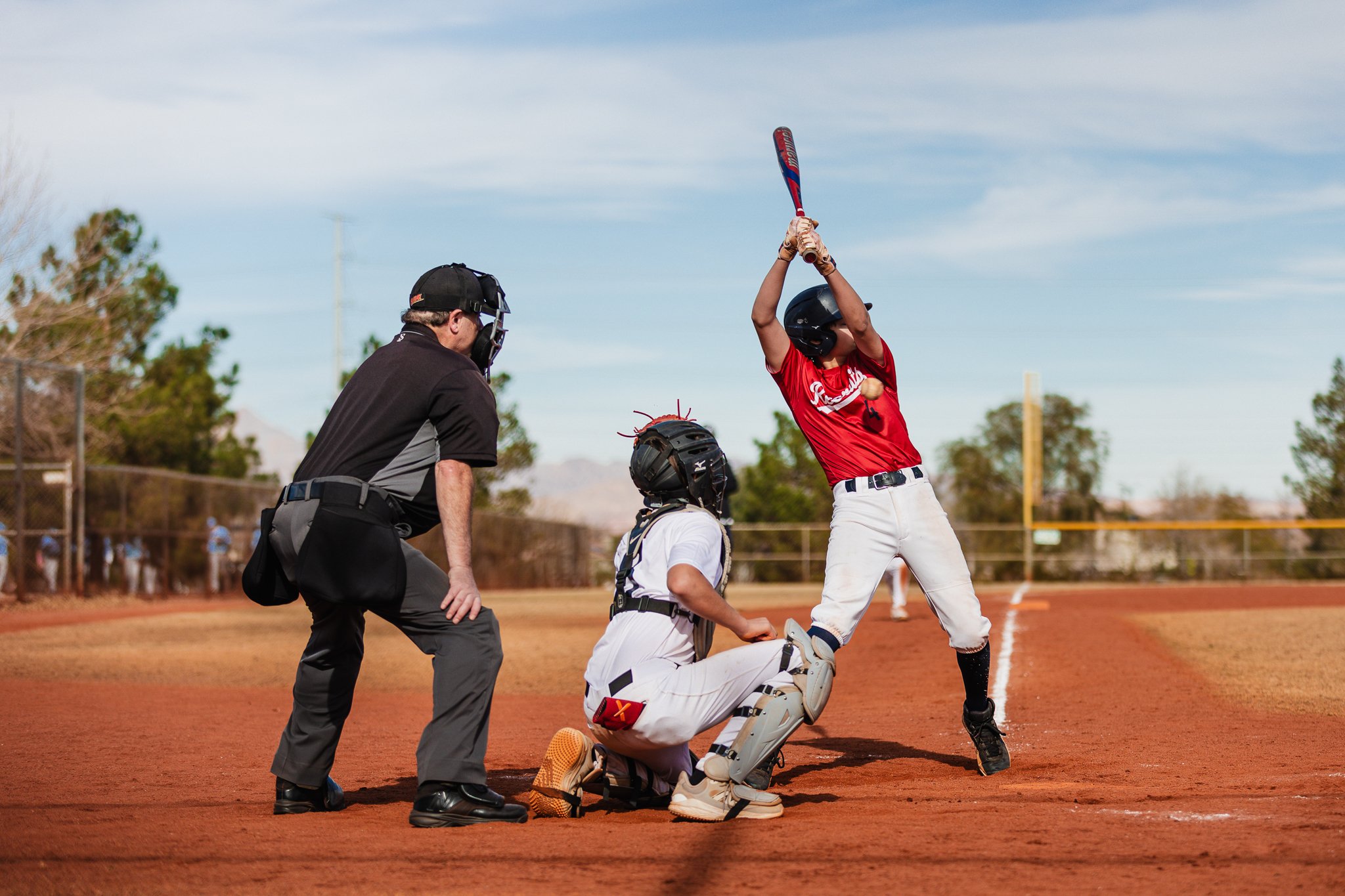 A baseball player in uniform standing at home plate preparing to bat, with an umpire, catcher, and a batter in a yellow helmet in position nearby during a game.