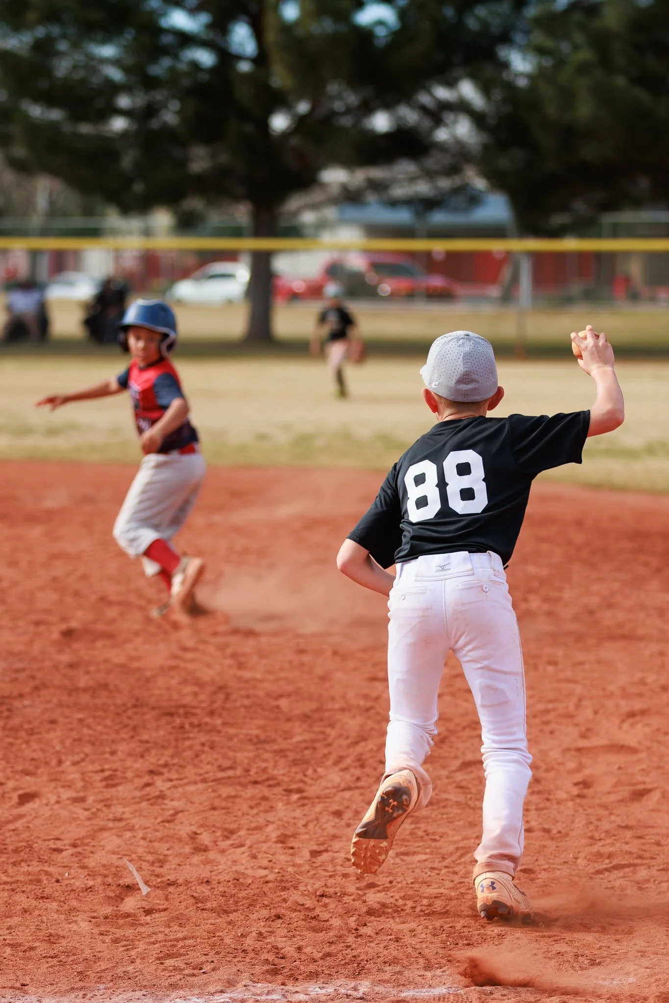 Young boys playing baseball on a field, with a player in a black jersey with the number 88 running, and a boy in a red jersey running behind him, with blurred background and trees.