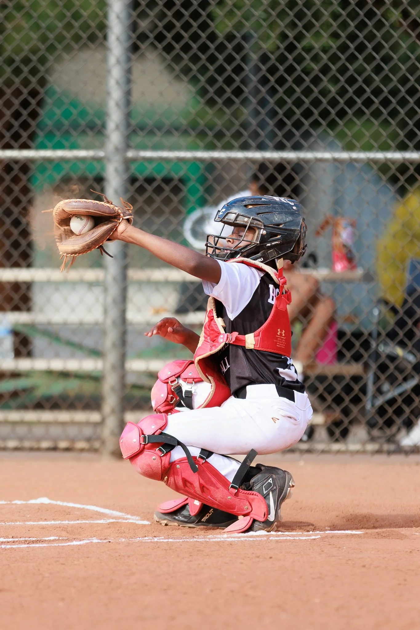 A young baseball catcher in a crouching position on a dirt field, wearing a black helmet with a face guard, a red and black protective chest guard, pink leg guards, and a white uniform, catching a baseball with a catcher's mitt.