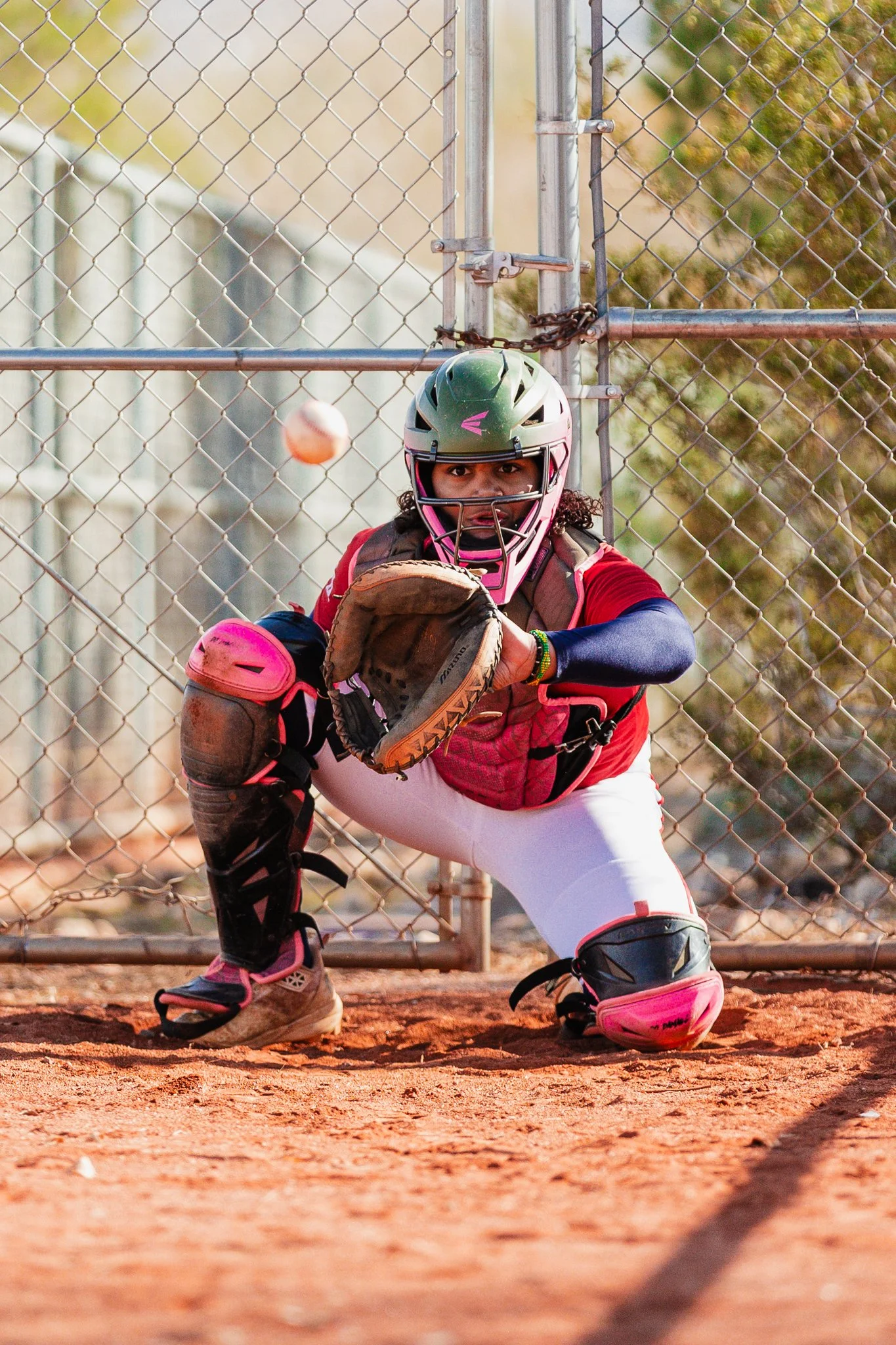 A female softball catcher in protective gear crouches behind home plate, ready to catch the incoming pitch. She is wearing a green helmet, a pink chest protector, and pink leg guards, with a glove positioned to catch the ball. The background shows a 