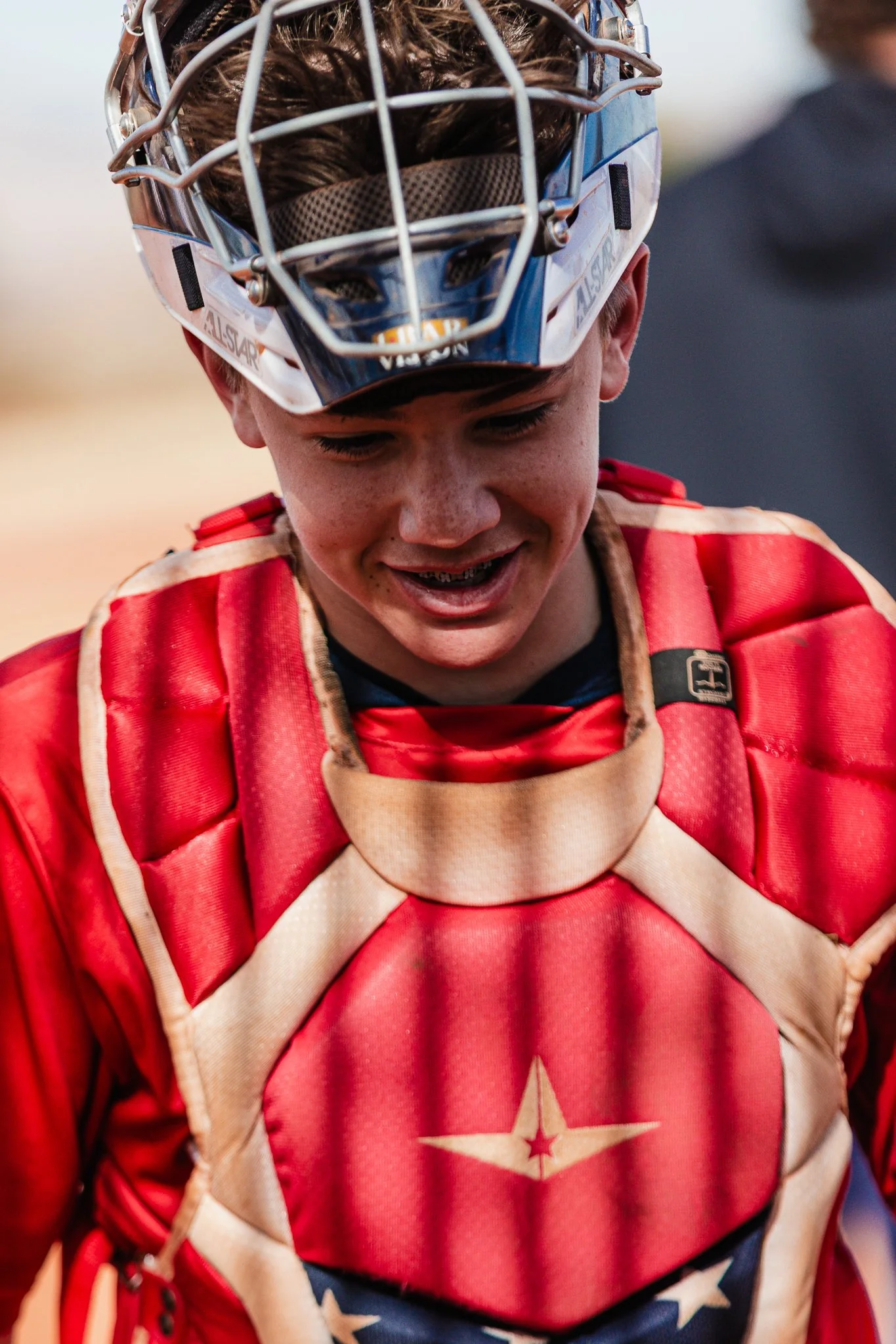 Close-up of a young hockey player wearing a red and beige team jersey with a star emblem, and protective gear including a helmet with a cage, as he looks downward.