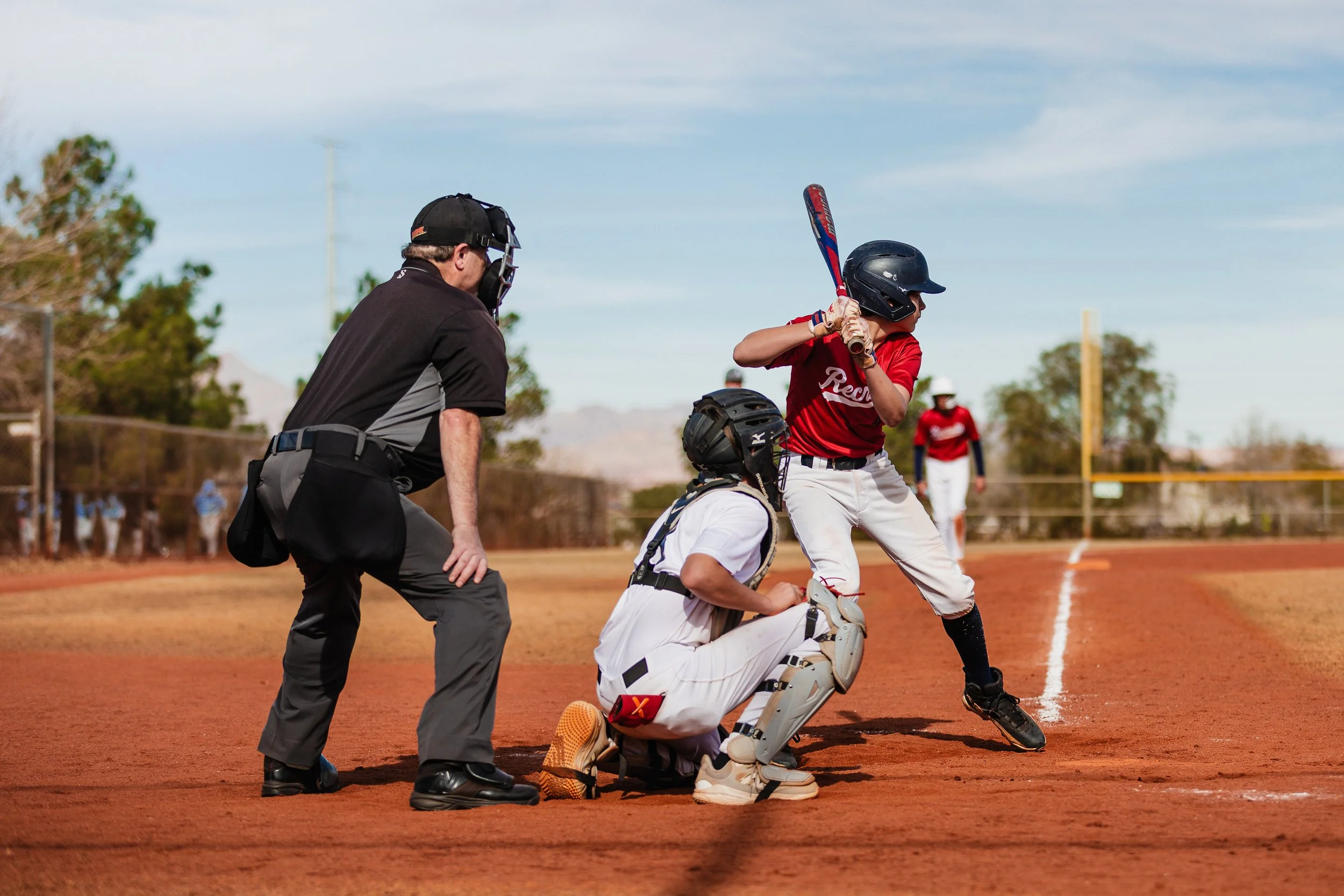 A baseball game scene showing a batter at home plate preparing to hit, a catcher and umpire crouched behind, and players in the background on a dirt field with a blue sky overhead.