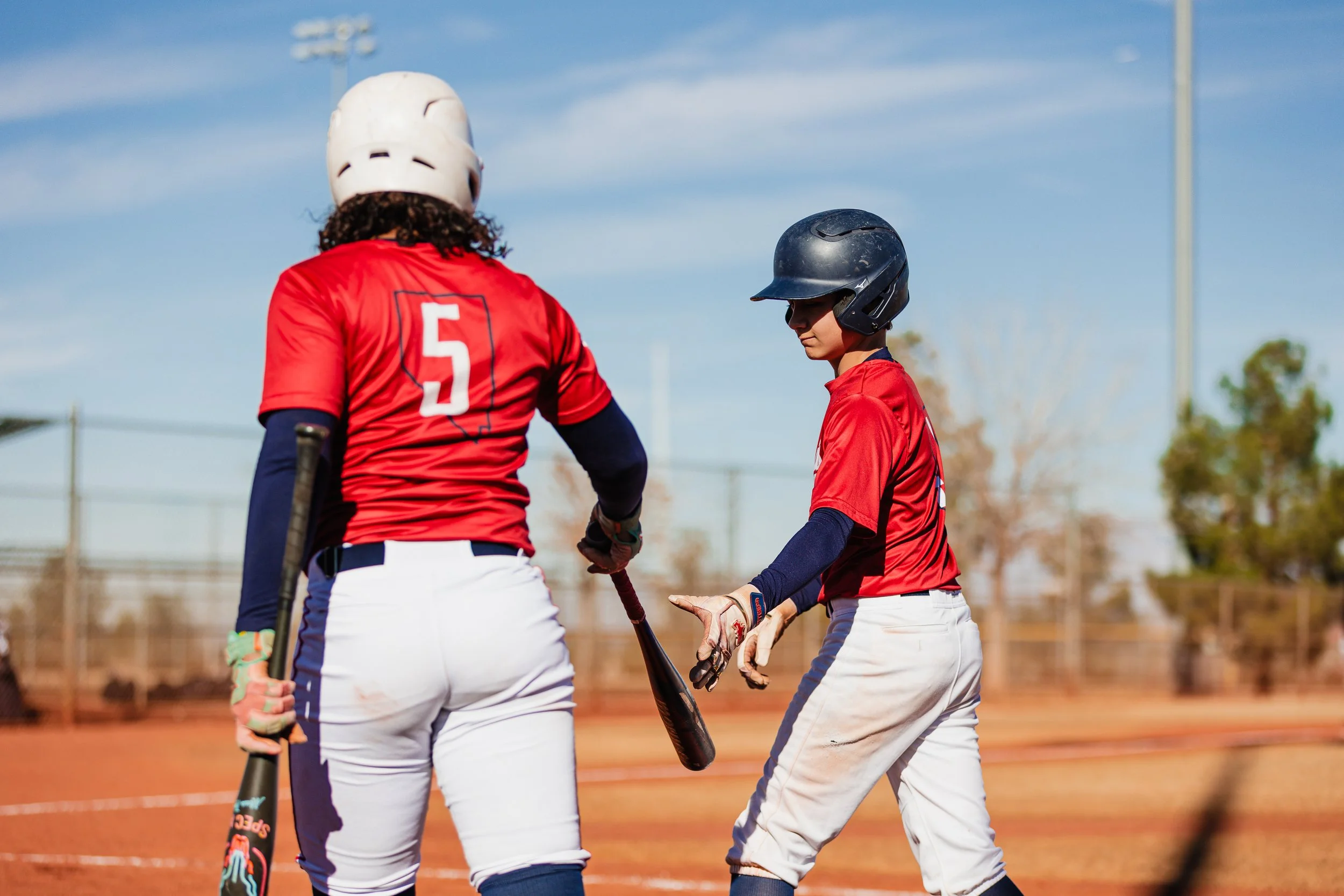 Two young baseball players in red uniforms and helmets stand on a baseball field, holding bats, during daytime with a clear blue sky.