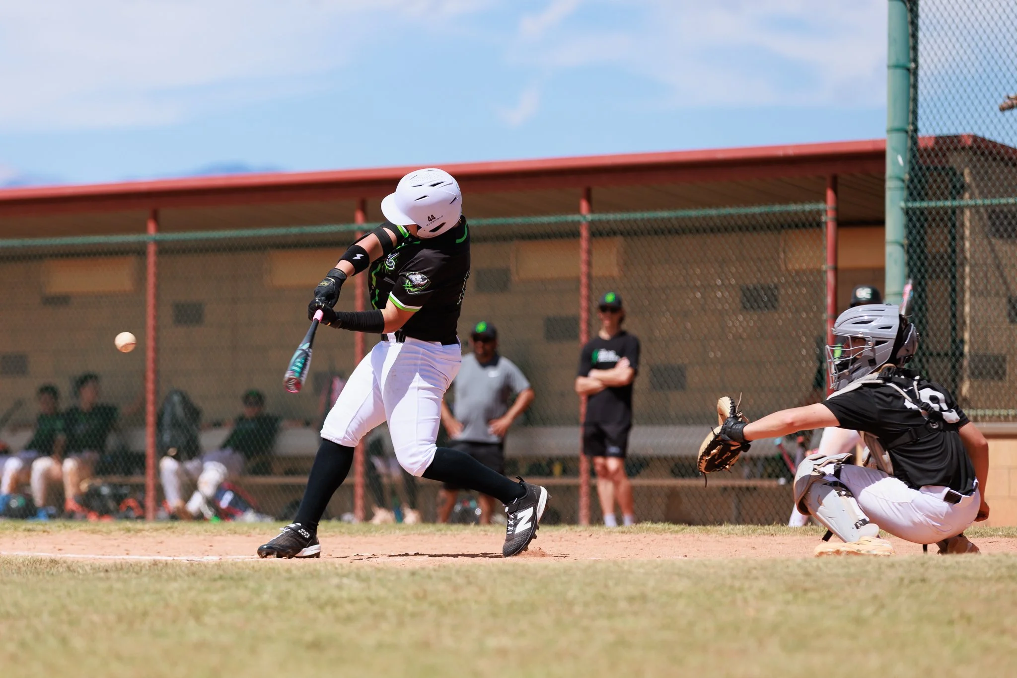 A baseball player swings a bat at a pitched ball during a game; the catcher is crouched behind home plate wearing a helmet and protective gear, and spectators watch from the sidelines.