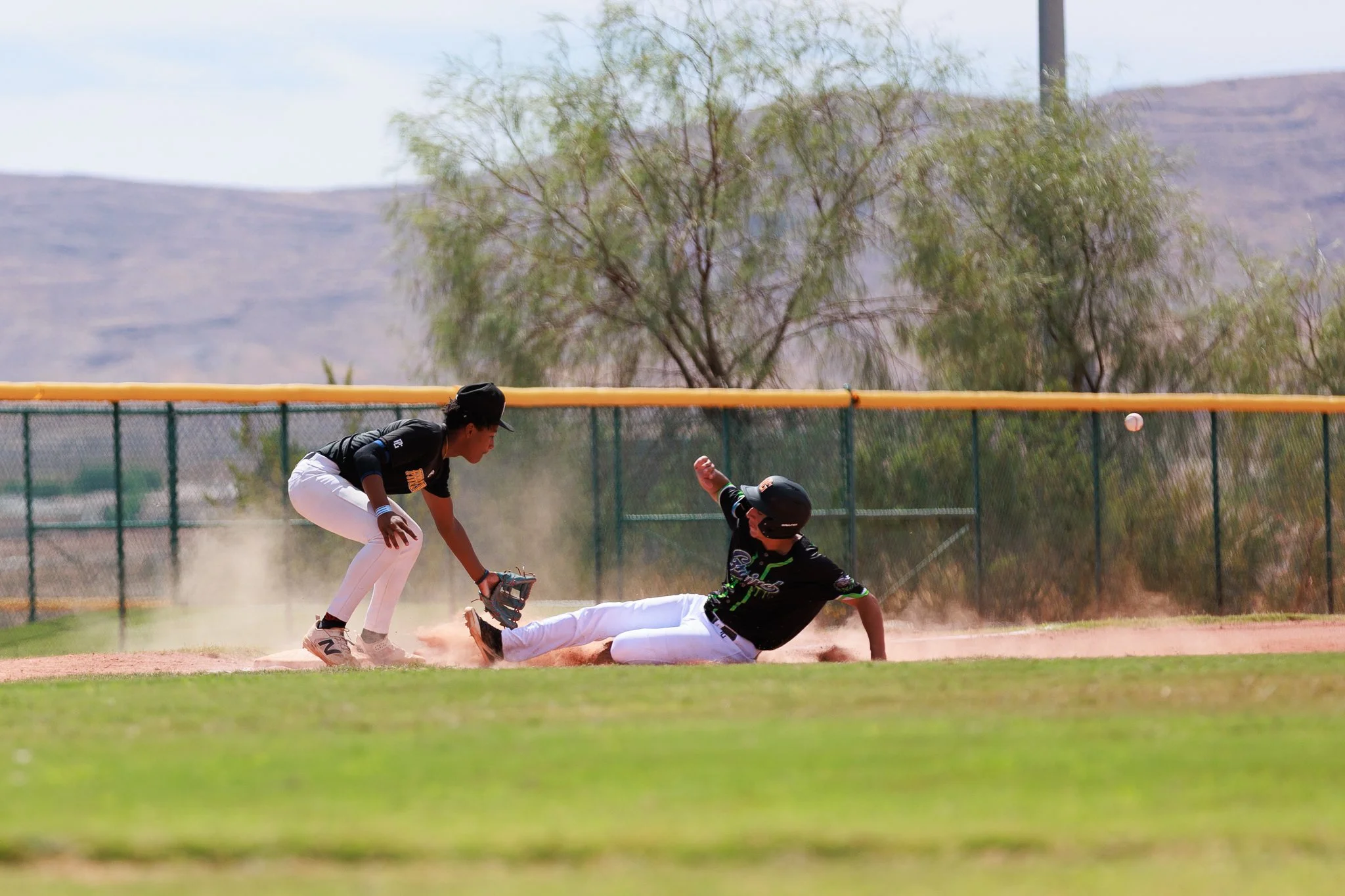 A baseball player slides into a base while two other players in black jerseys and a player in a black and green jersey attempt to tag him out on a baseball field, with a fence and trees in the background.