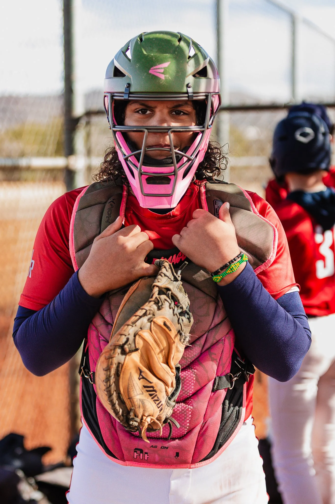 Close-up of a female baseball player wearing a catcher's helmet and gear, holding a baseball glove, with other players in background.