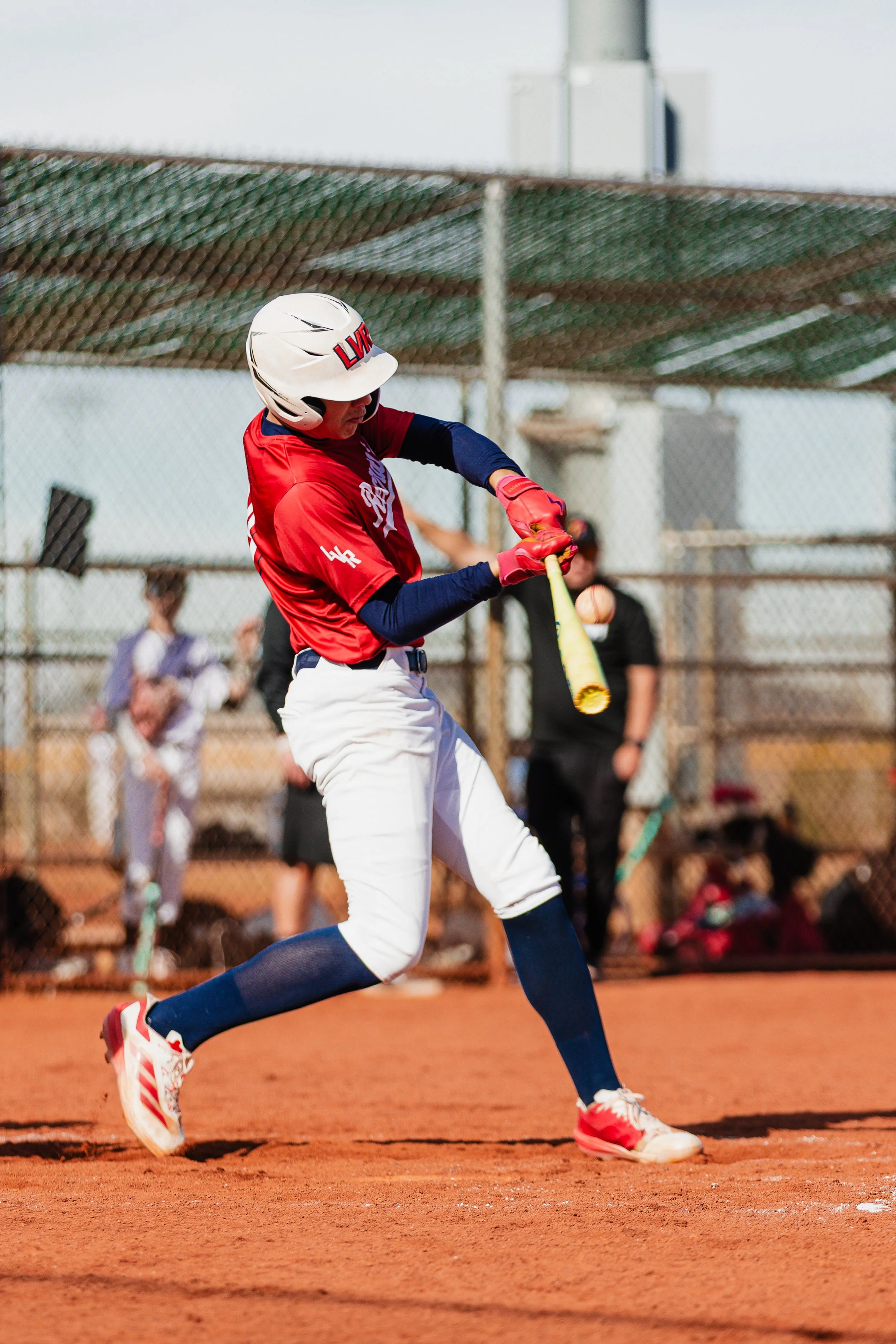 A baseball player in red and white uniform is hitting a ball with a yellow bat on a baseball field with a chain-link fence in the background.