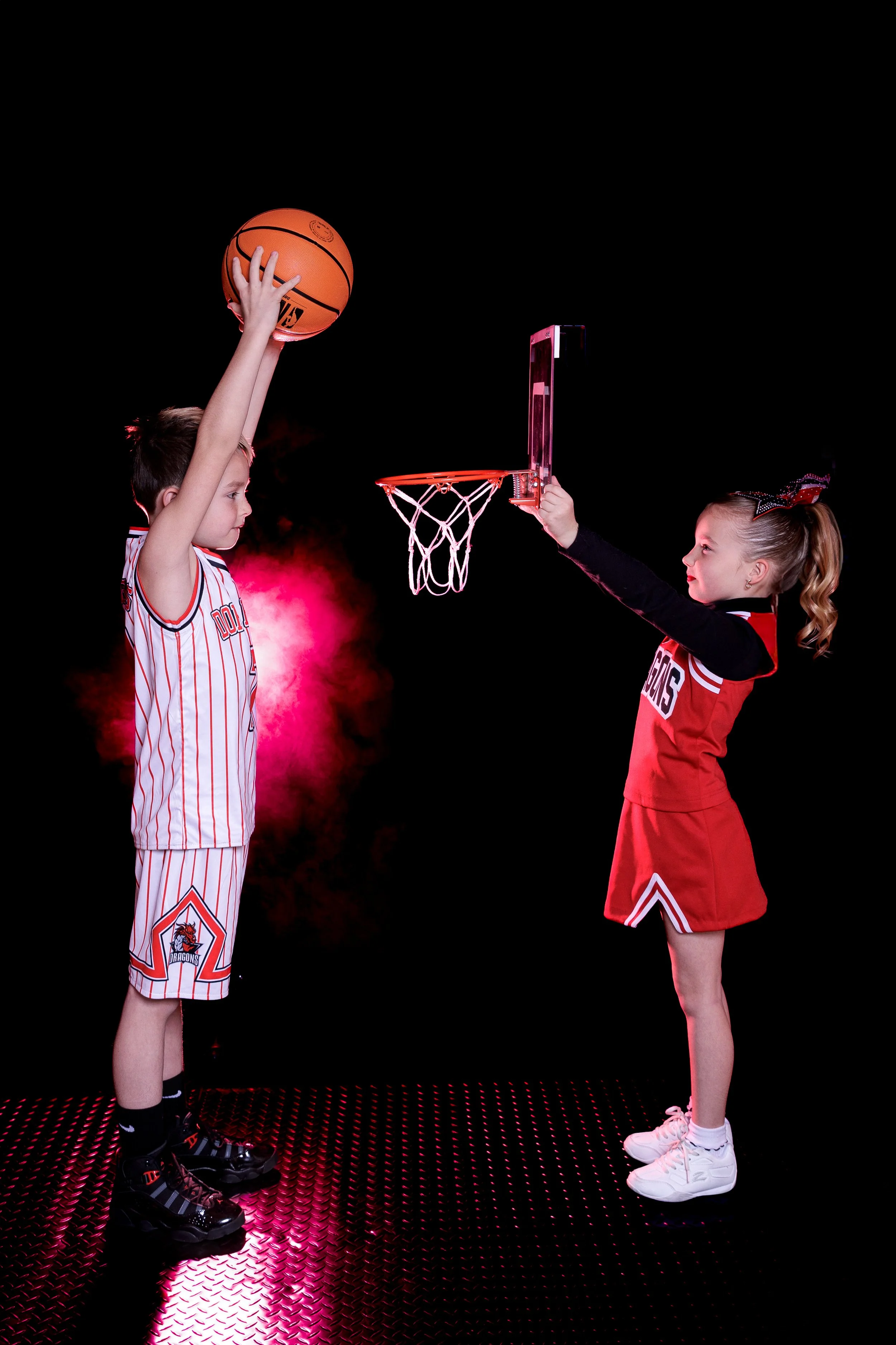 Two young children, a boy and a girl, playing basketball in a studio with a black background and pink lighting. The boy is about to take a shot, and the girl is holding a small basketball hoop, as if about to make a shot herself. The boy wears a white and red basketball uniform and the girl wears a red and black basketball uniform. Both children are standing on a textured, rubberized floor.