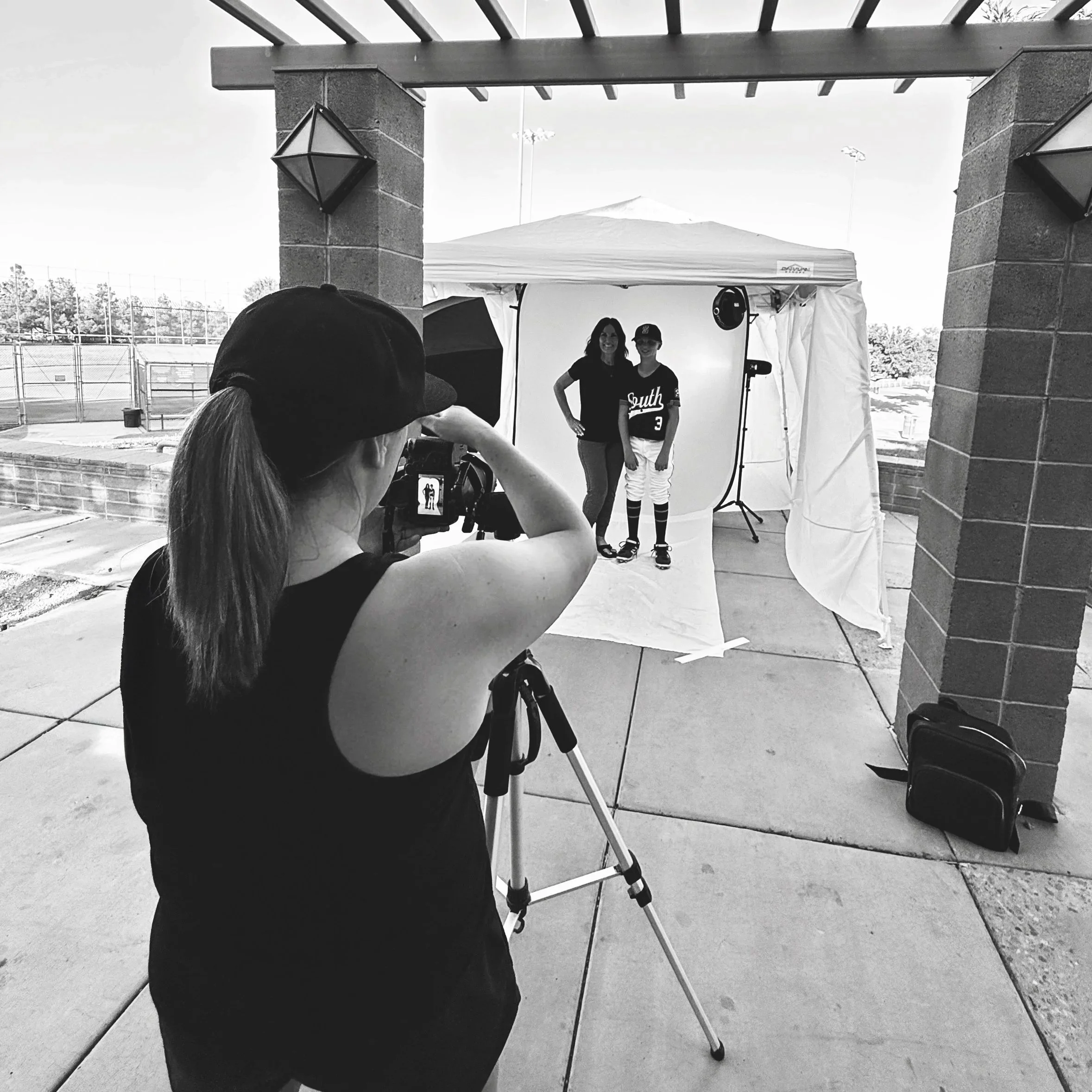 Two women standing side by side in front of a photography backdrop at an outdoor event, with a person in the foreground taking photos. The women are smiling, and one is wearing a baseball jersey with the word 'South' and the number 3 on it.