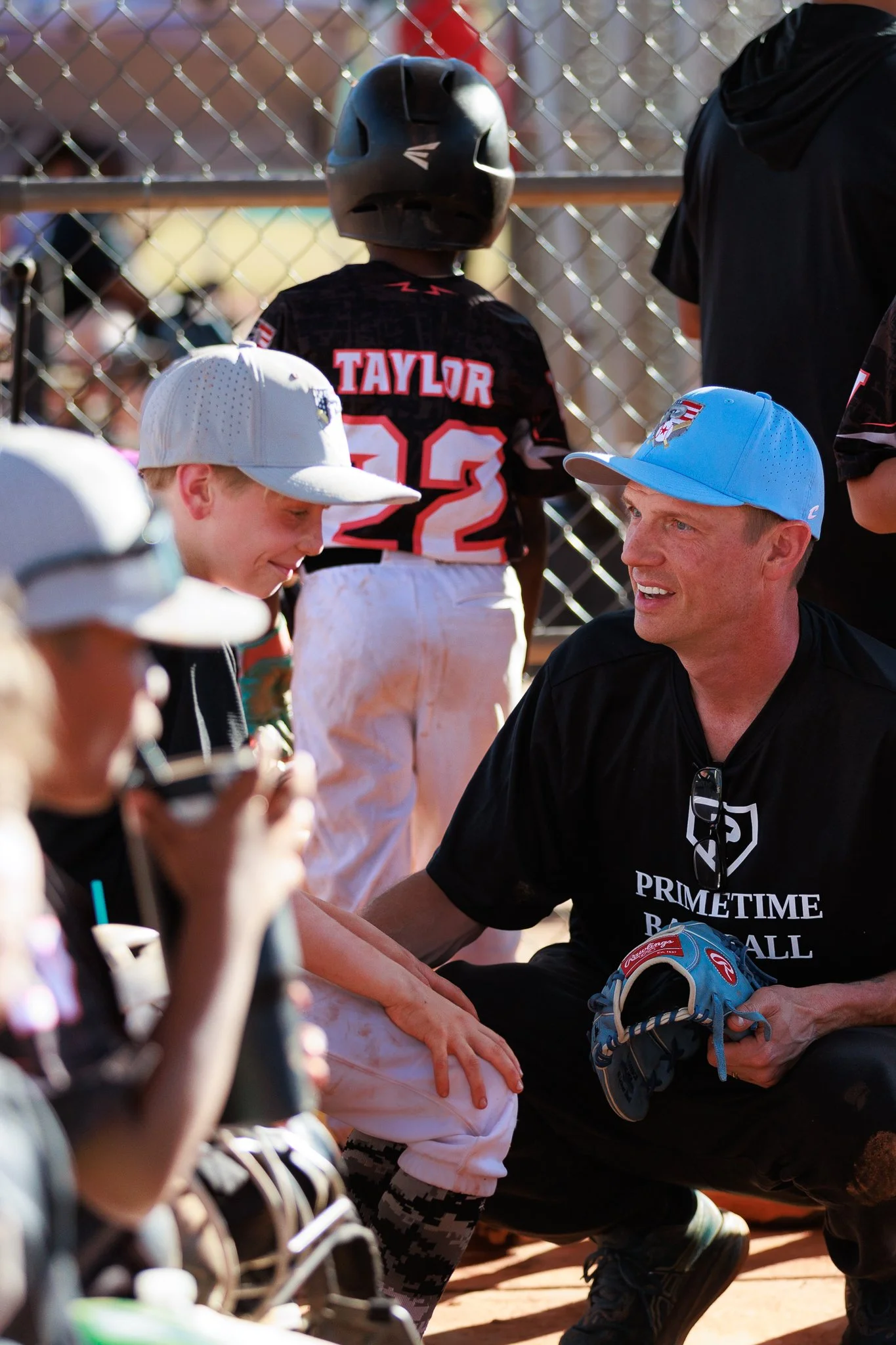 A man in a black shirt and cap interacting with a young boy wearing a white baseball cap at a baseball game, with players and a chain-link fence in the background.
