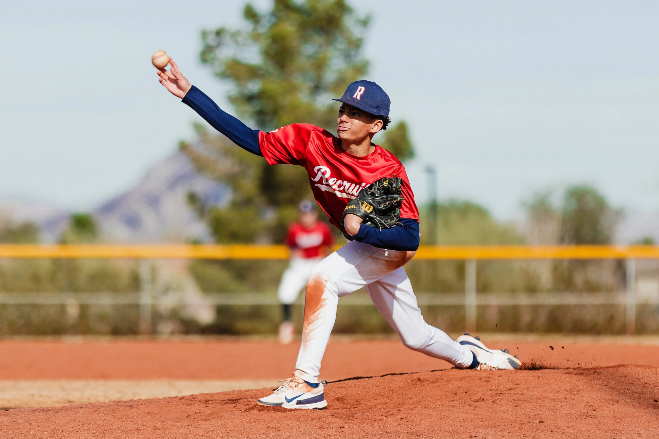 A young baseball player wearing a red and navy uniform is fielding a ground ball on the infield, with his glove reaching down and his body in a low, side-on stance. The player is wearing a navy cap with a red and white letter, and the background features grassy outfield with trees and other players in the distance.
