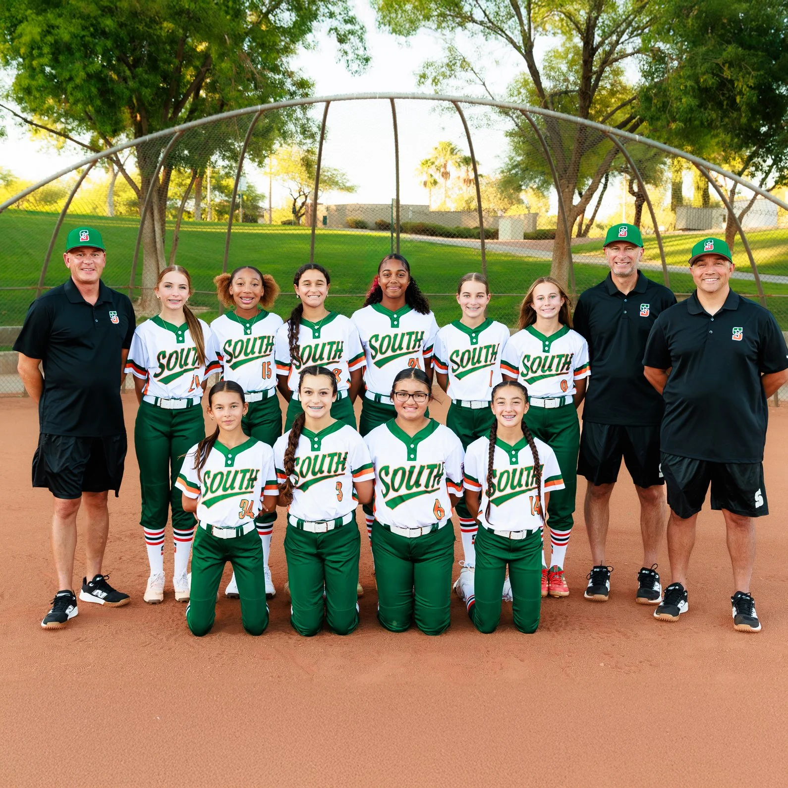 A youth softball team with players and coaches posing in two rows on a softball field, under trees in a park during daytime.