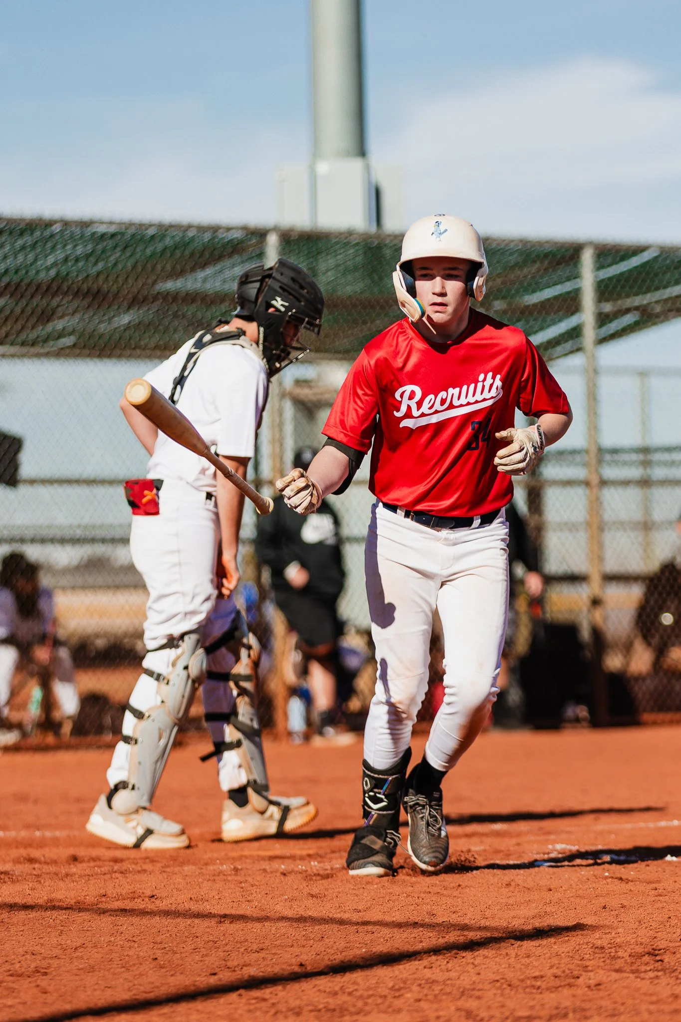 Two baseball players on the field, one in catcher's gear and the other in a red jersey with 'Recruits' written on it, interacting during a game or practice on a sunny day.