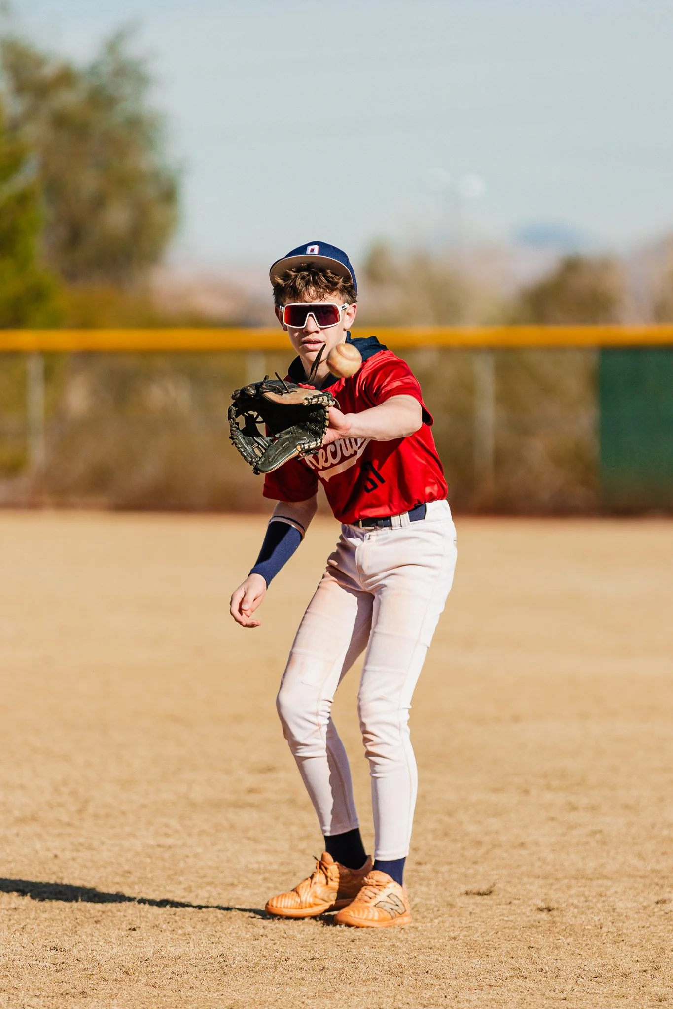 A young baseball player in a red jersey, white pants, wearing sunglasses and a baseball cap, is on the field preparing to catch a ball with his glove.