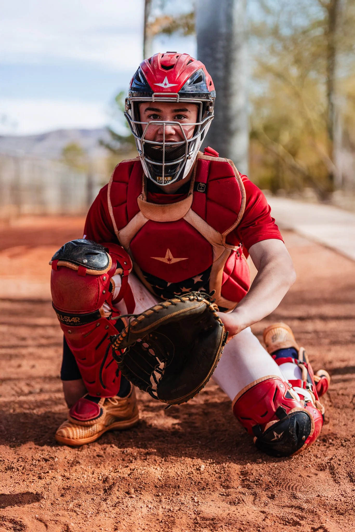 A young baseball catcher in a red uniform and protective gear, crouching on a dirt field during daytime, ready to catch a ball.