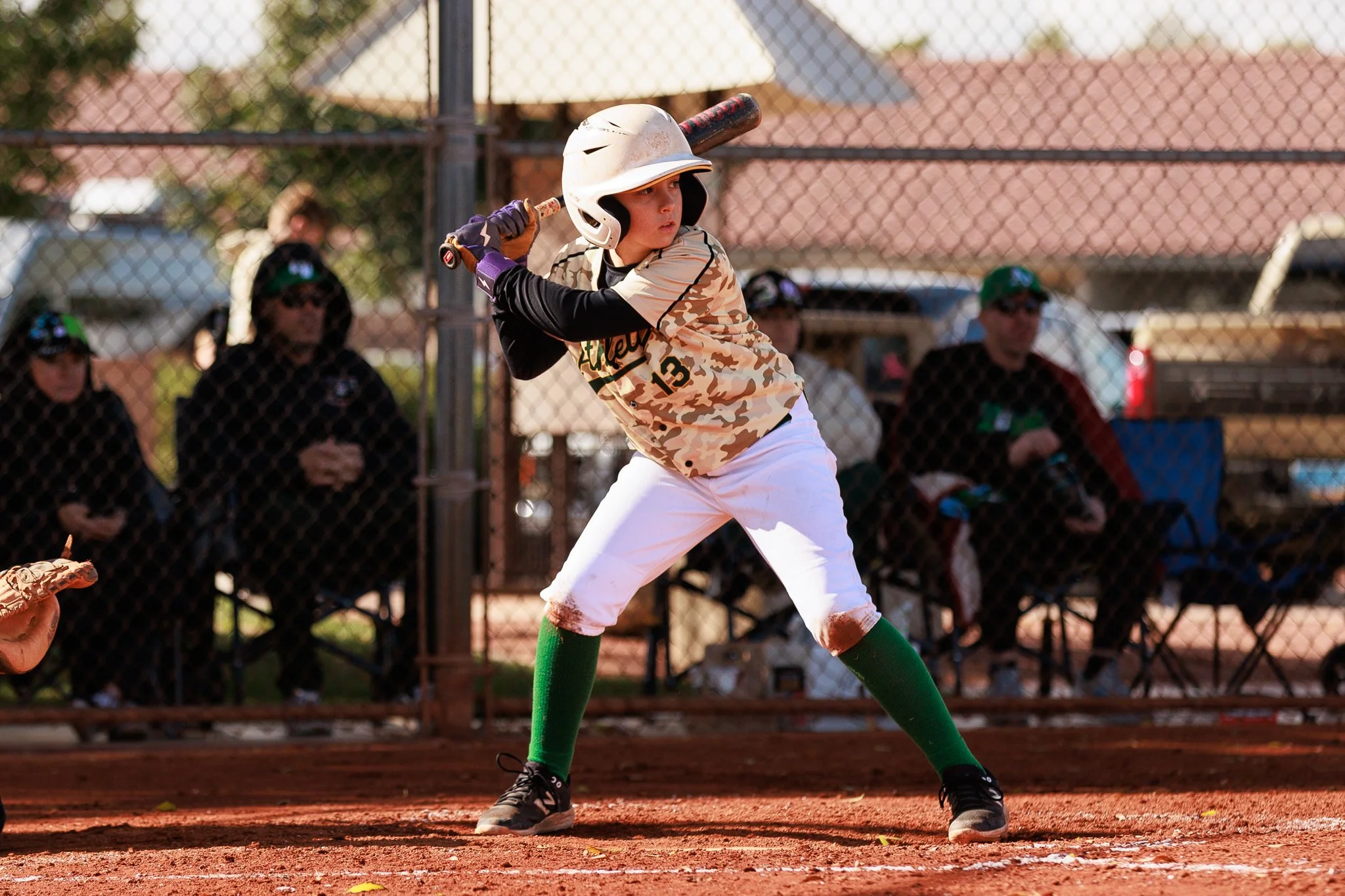 A young baseball player at bat, wearing a white helmet and camouflage jersey, swinging at a pitched ball on a baseball field.