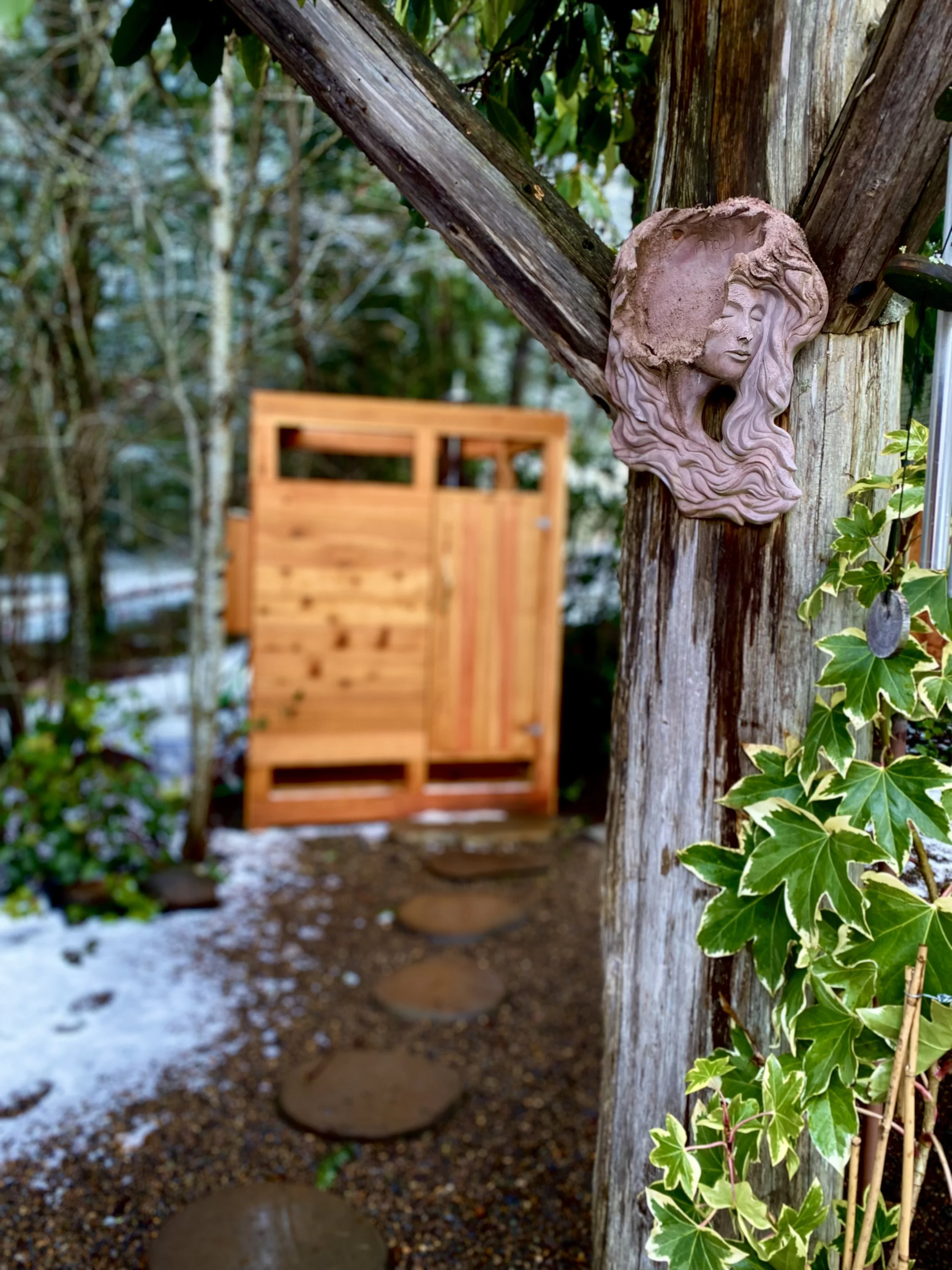 Close-up of a decorative wall sculpture of a woman's face with flowing hair, mounted on a weathered wooden post in an outdoor garden. A wooden gate and stepping stones are visible in the background, with green foliage and some snow on the ground.