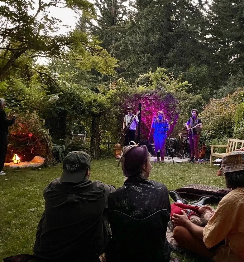 A live outdoor musical performance at dusk in a wooded backyard, with three musicians on stage and an audience sitting on the grass.