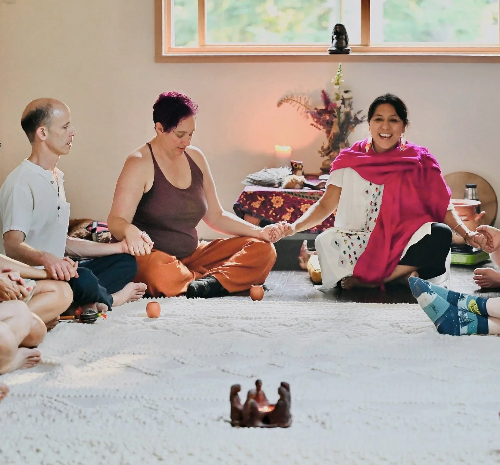 People participating in a spiritual or religious ceremony sitting on the floor in a circle, holding hands, with a window and decorative items in the background.