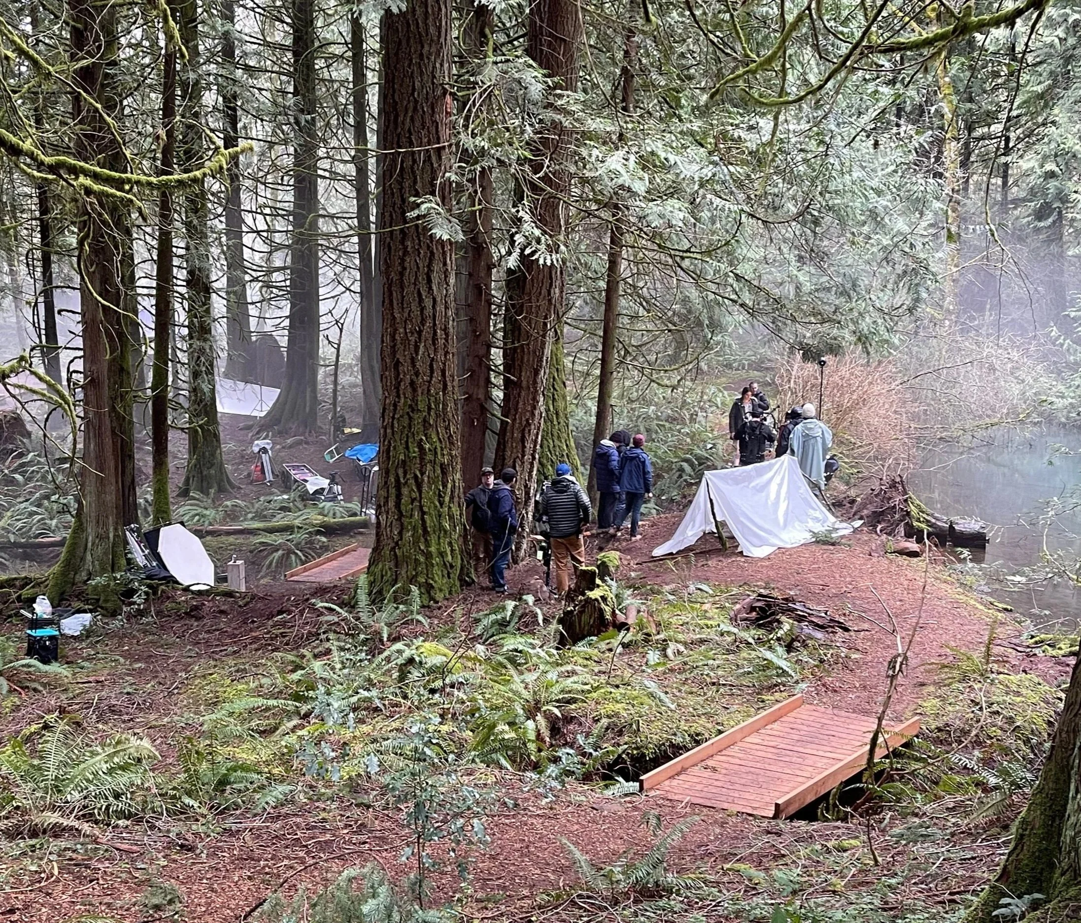 Film or photography crew working in a forest, with people setting up equipment, a white tent, and wooden pathway in a dense, misty forested area.