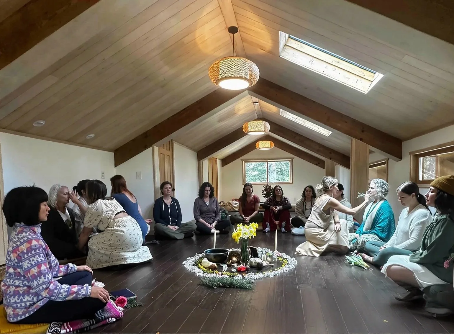 Group of women participating in a spiritual or wellness ceremony in a cozy, wooden-floored attic room with skylights, sitting in a circle around a decorated altar with flowers, candles, and stones.
